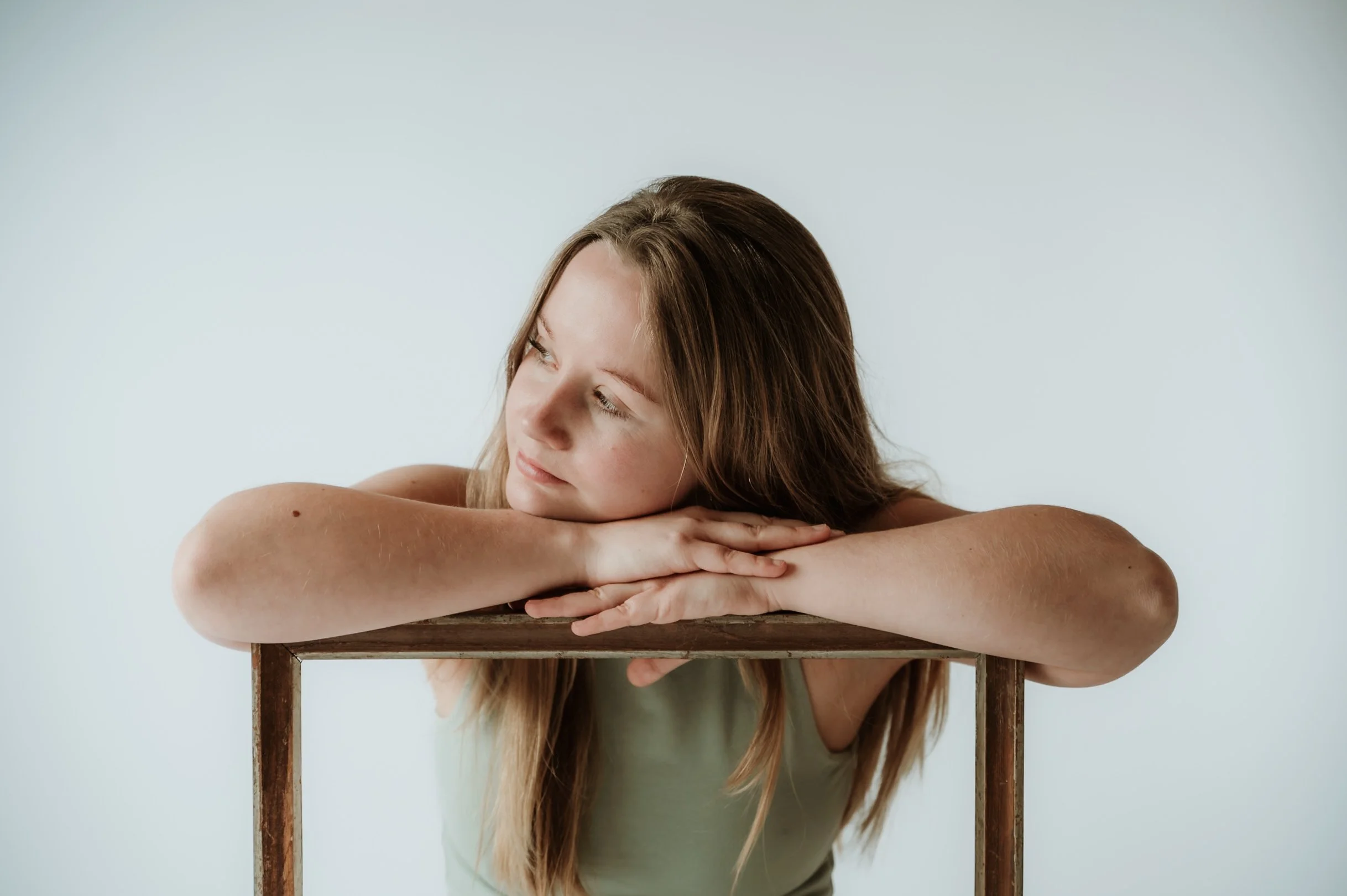 A woman with long brown hair leaning her head on her arms, which are resting on a wooden table, looking to the side against a plain light background.