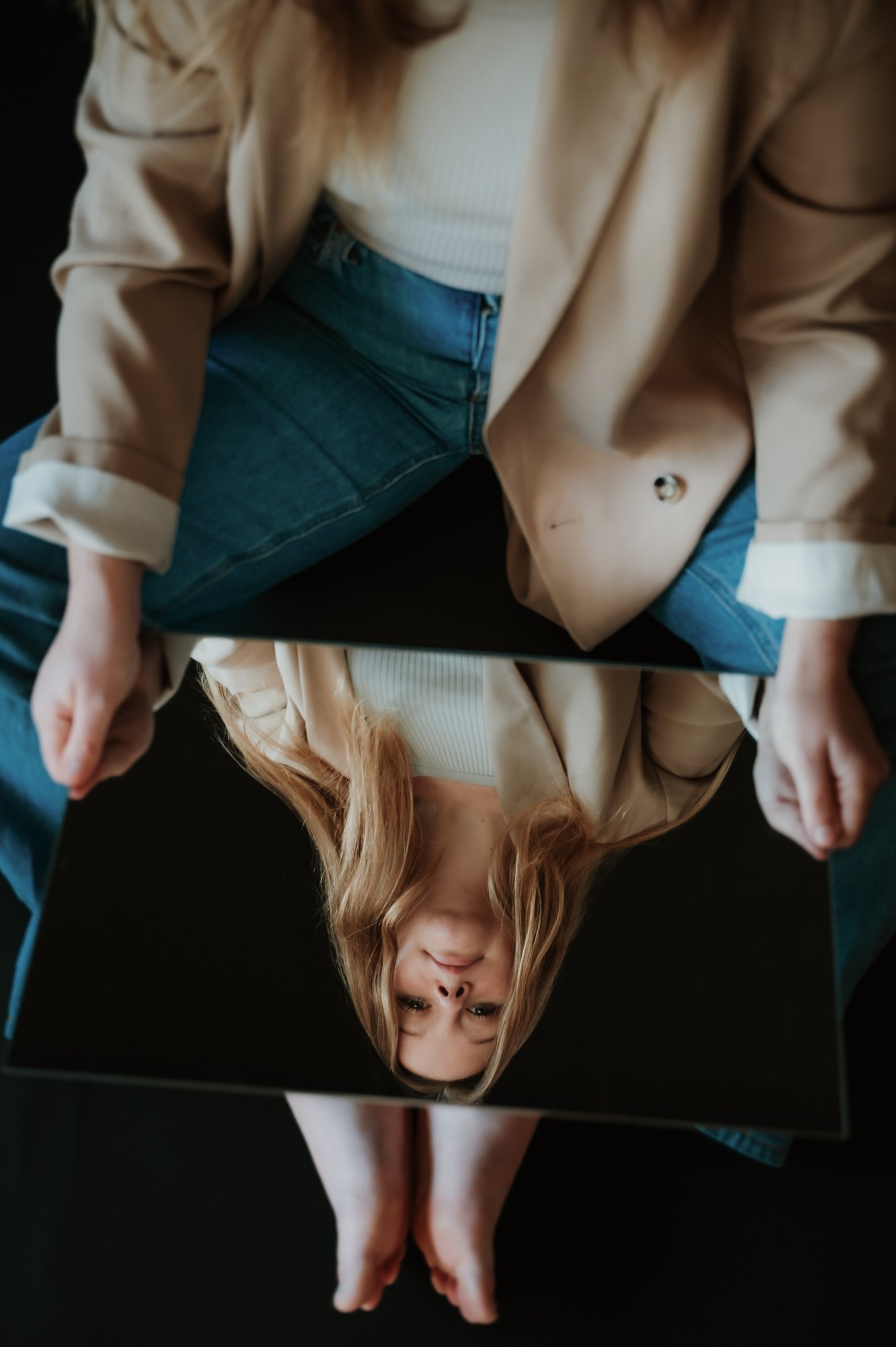 A woman with long, wavy blonde hair is lying on her back on a black surface. She is wearing a beige jacket, a white ribbed top, and blue jeans. Her head is tilted downward and her face is partially visible, with her eyes closed and a slight smile. Th