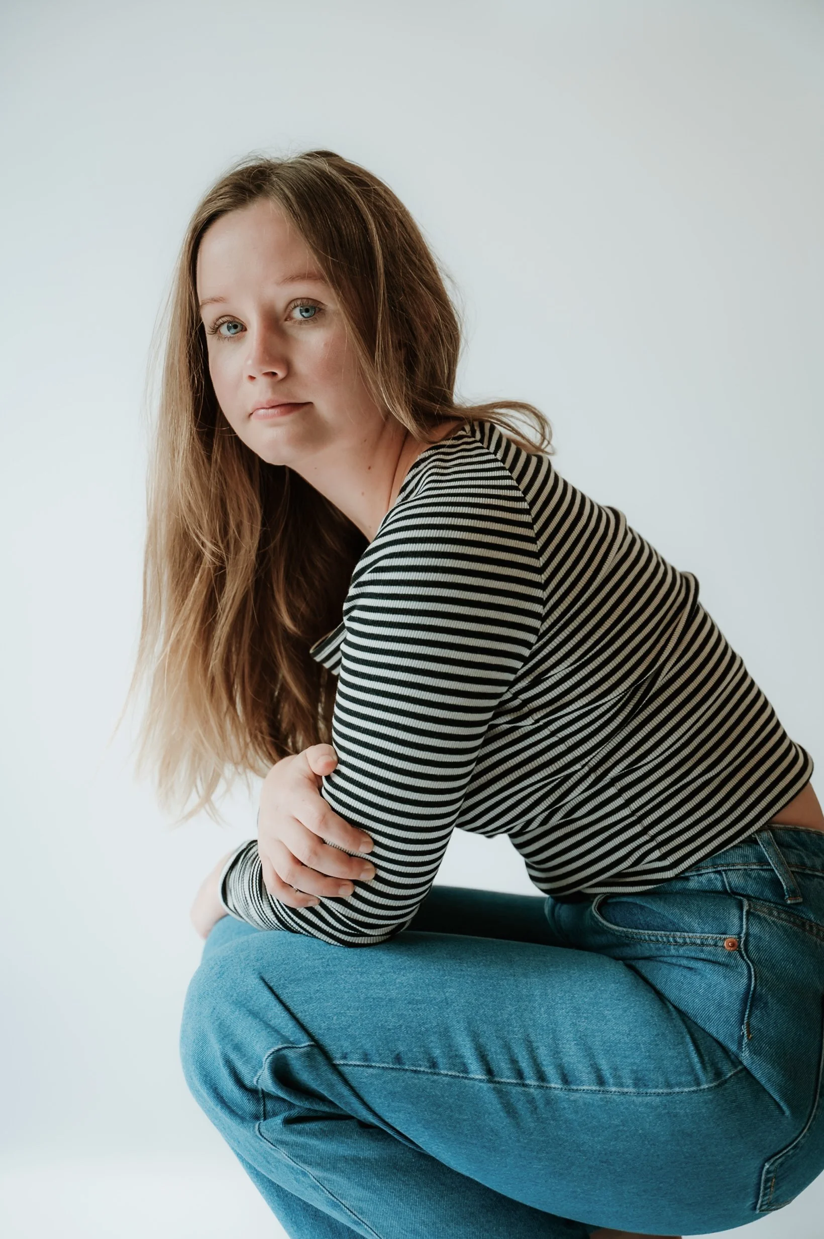 A young woman with long, wavy hair wearing a striped long-sleeve shirt and jeans, sitting on a chair with her arms crossed over her knees in a minimal, light-colored room.