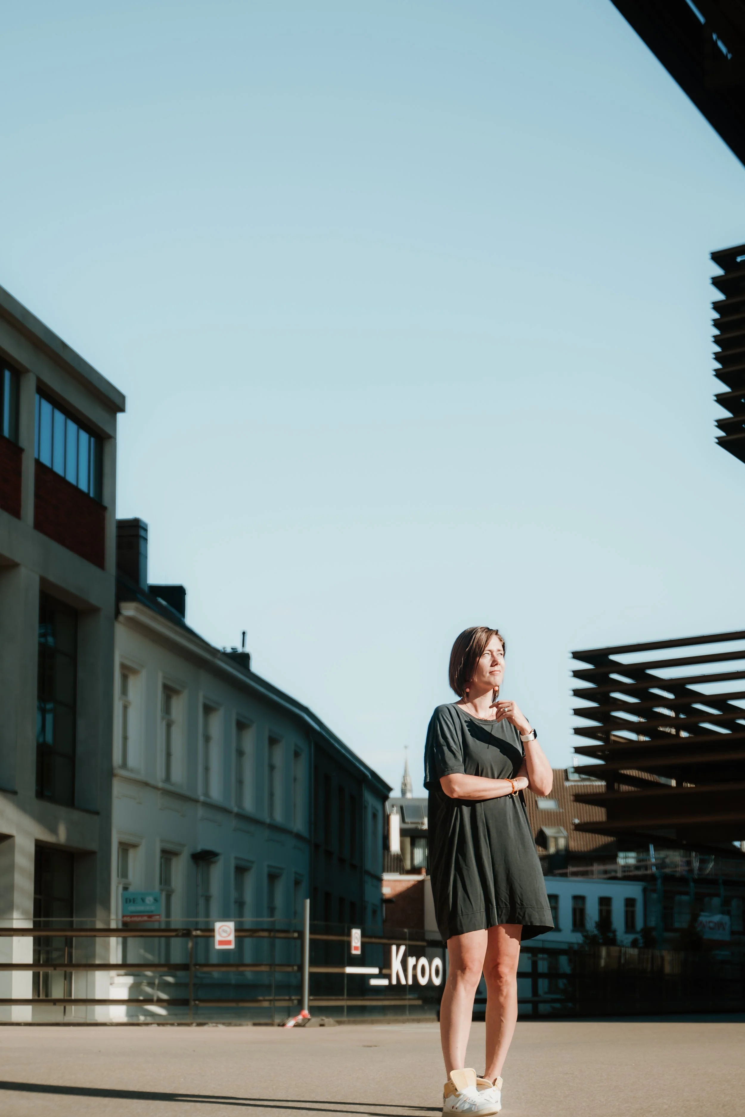 A woman with short hair dressed in a black oversized t-shirt and shorts stands outside on a sunny day, surrounded by modern buildings and a rooftop structure, with a clear blue sky overhead.