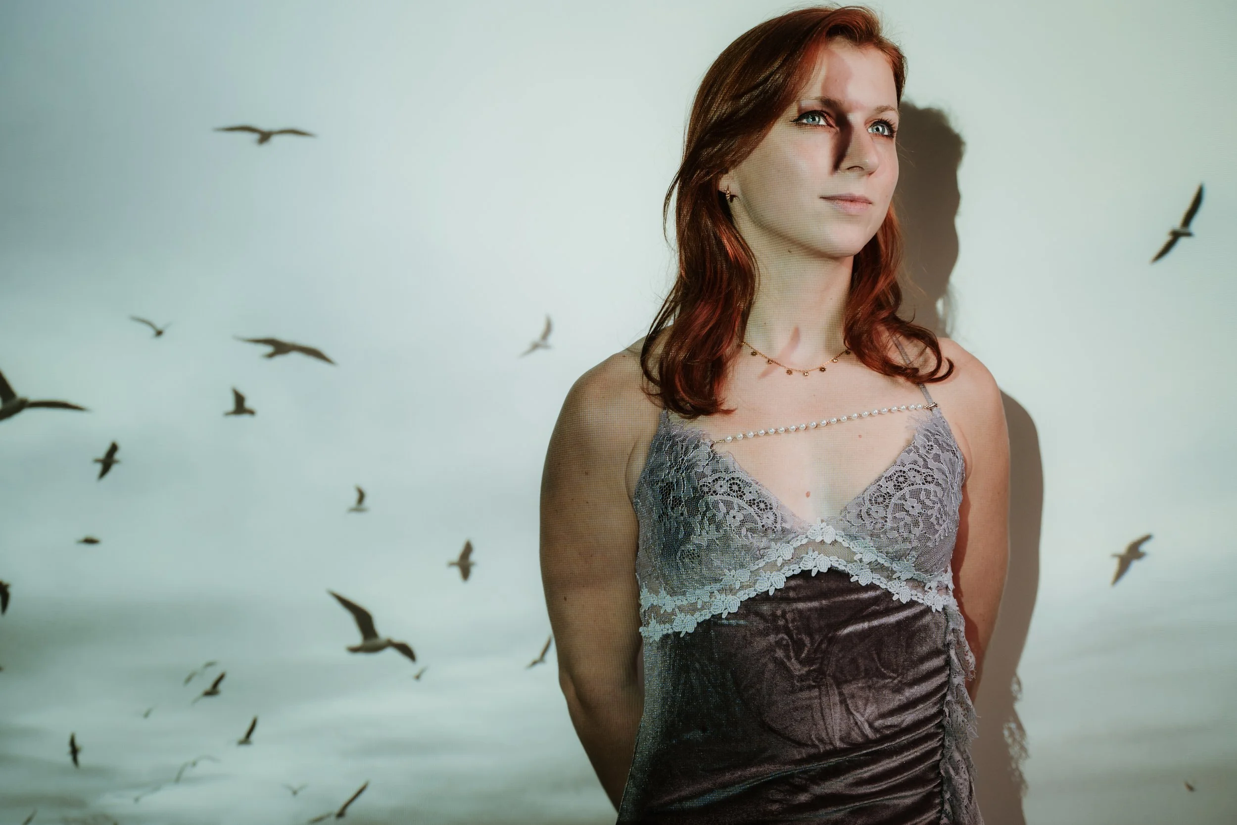 A woman with red hair in a gray lace and satin dress standing against a white background with silhouettes of flying birds.