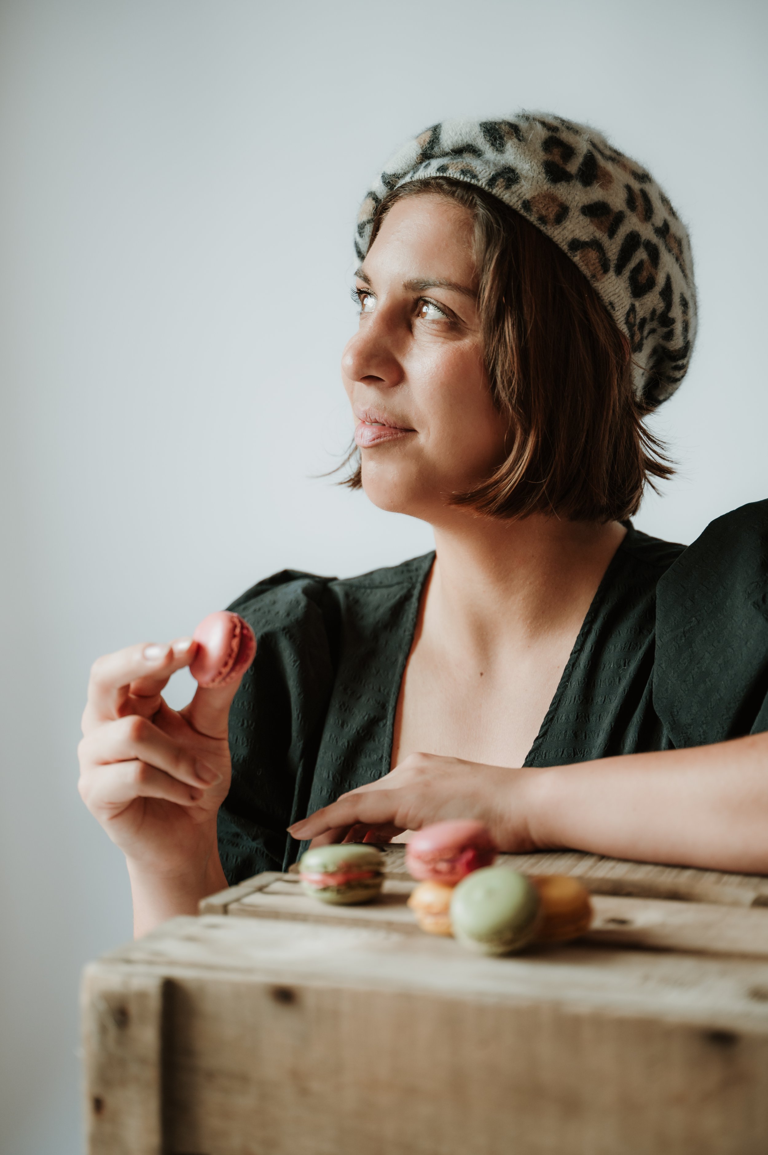 A woman with short brown hair wearing a leopard print beret, holding a pink macaron, and sitting at a wooden table with colorful macarons and pastries.