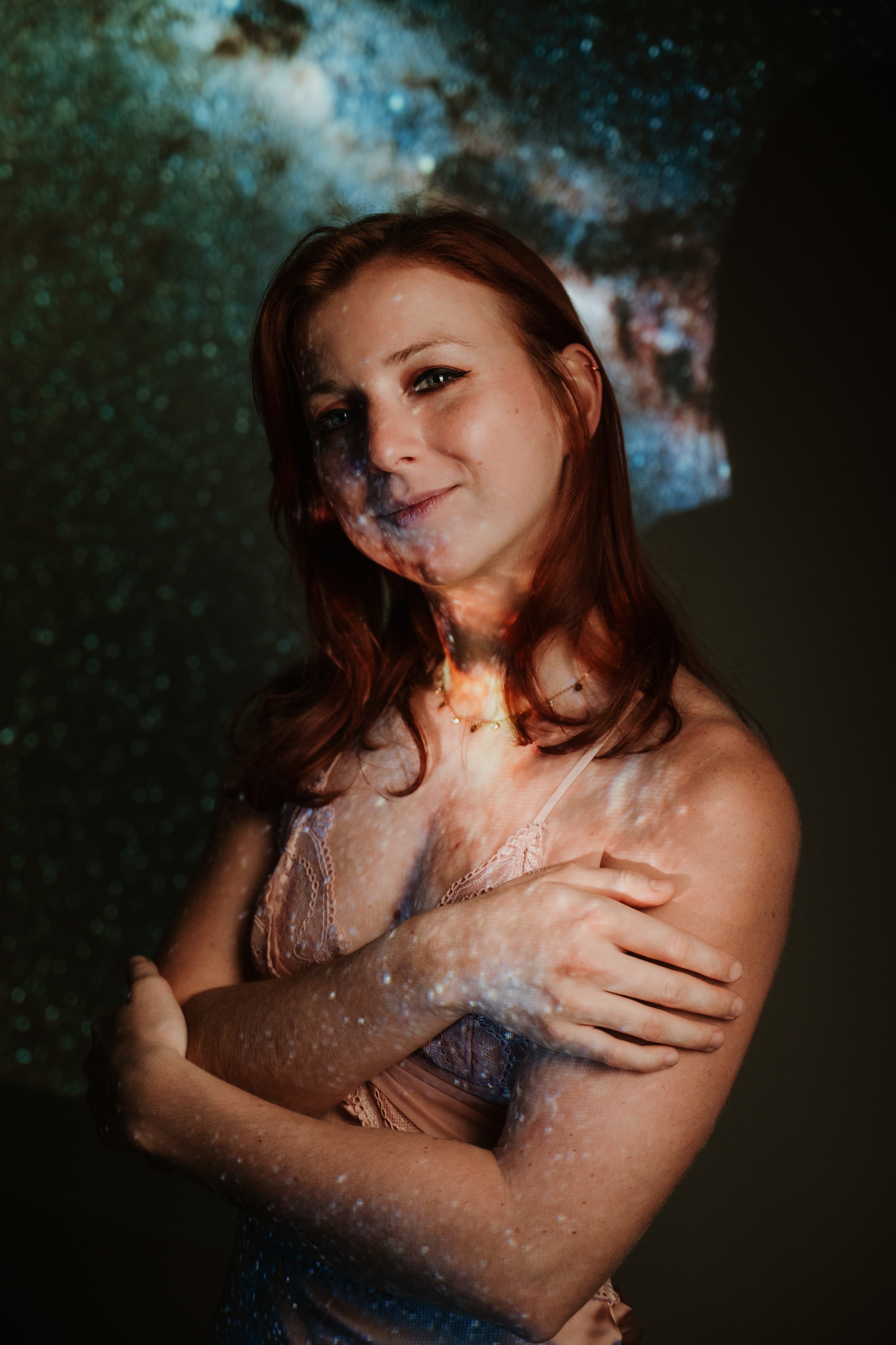A young woman with auburn hair and blue eyes standing with her arms crossed, illuminated by multicolored galaxy-like light patterns projected onto her face and body against a dark background.
