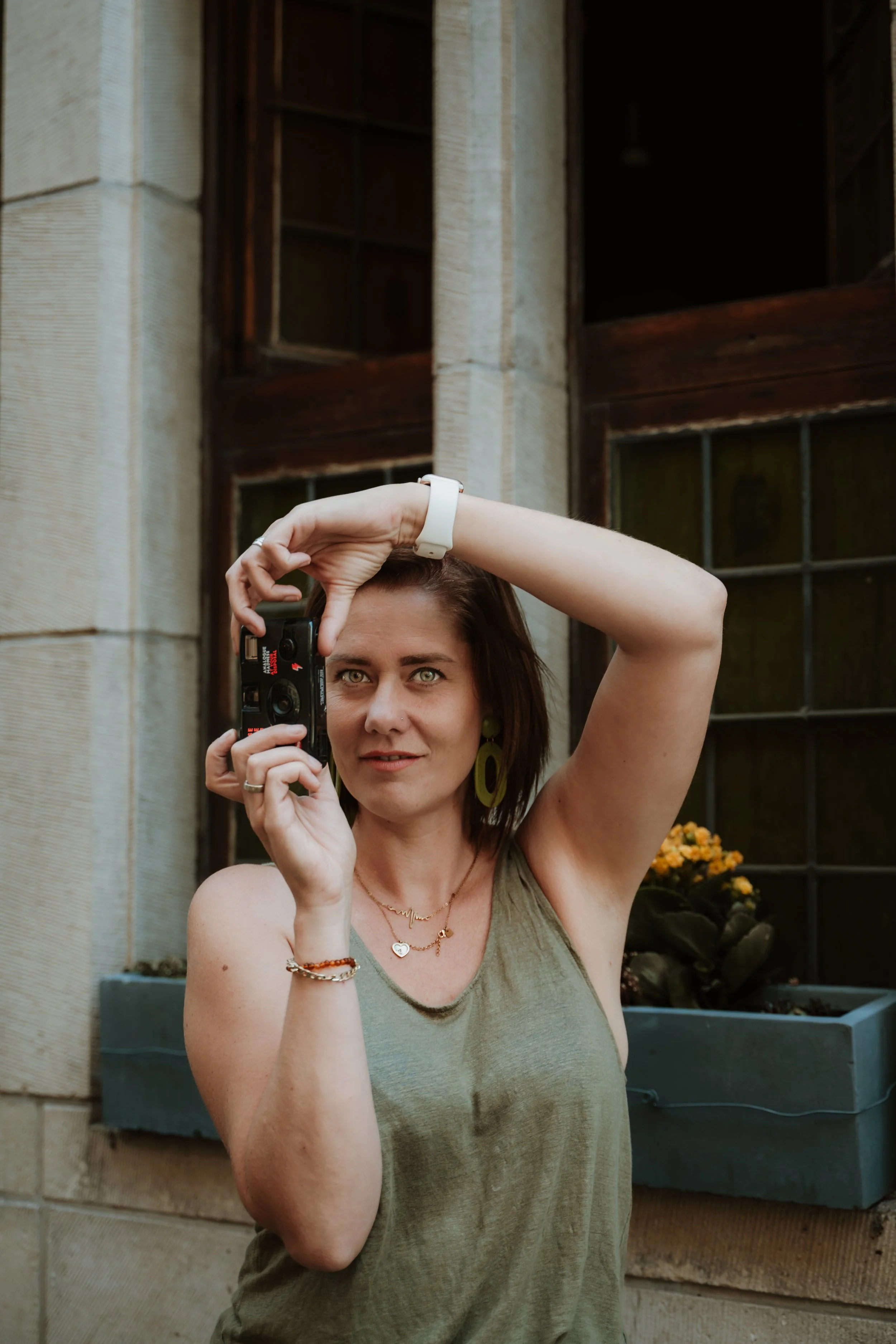 A woman with shoulder-length brown hair and light-colored eyes standing outside, taking a selfie with a camera. She is wearing a sleeveless olive-green top, a white watch, earrings, and layered necklaces, with a reddish bracelet on her wrist. Behind 