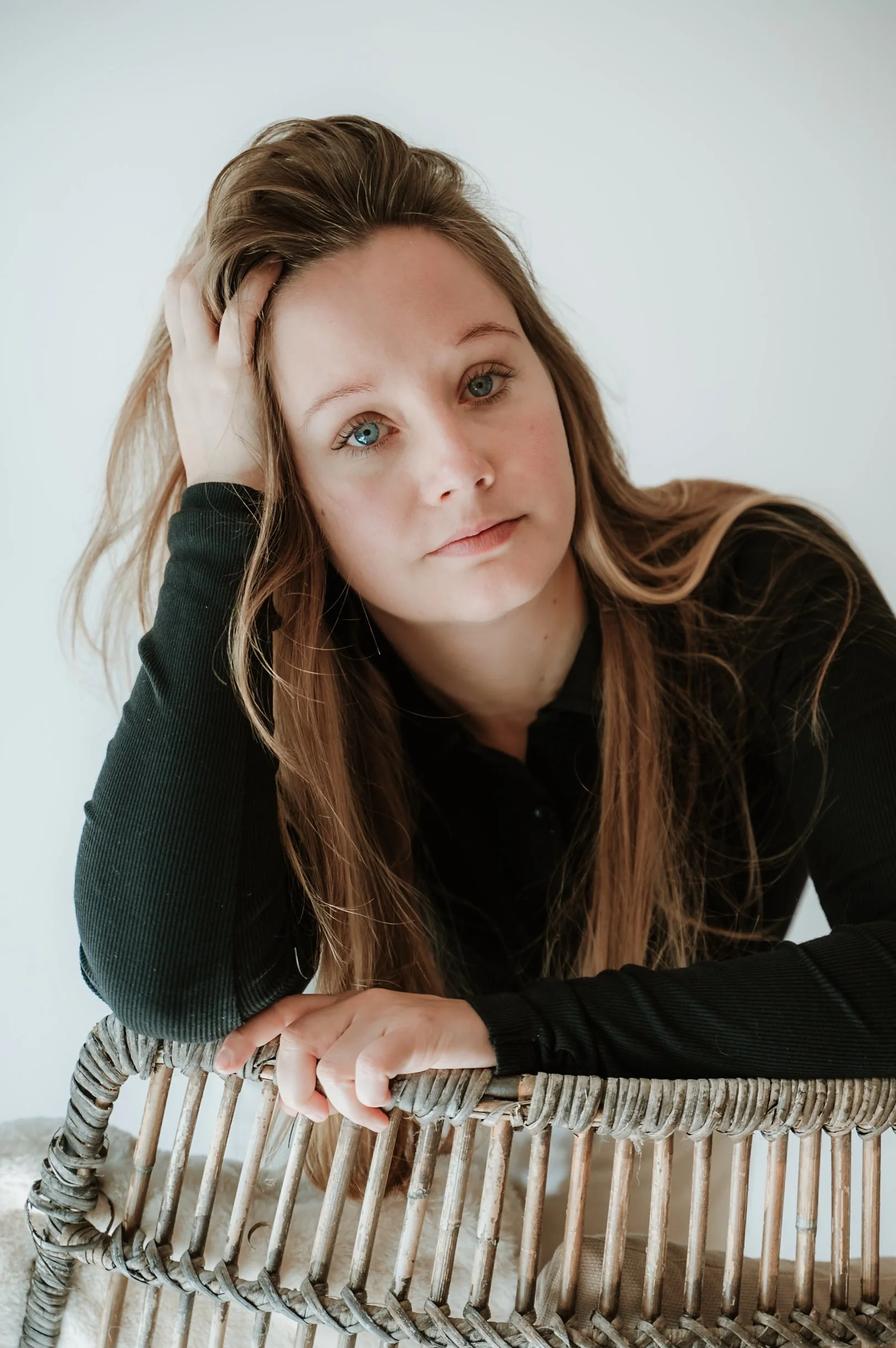 A woman with long, light brown hair and blue eyes, wearing a black long-sleeve shirt, rests her head on her hand while leaning on a wicker chair, with a neutral wall background.
