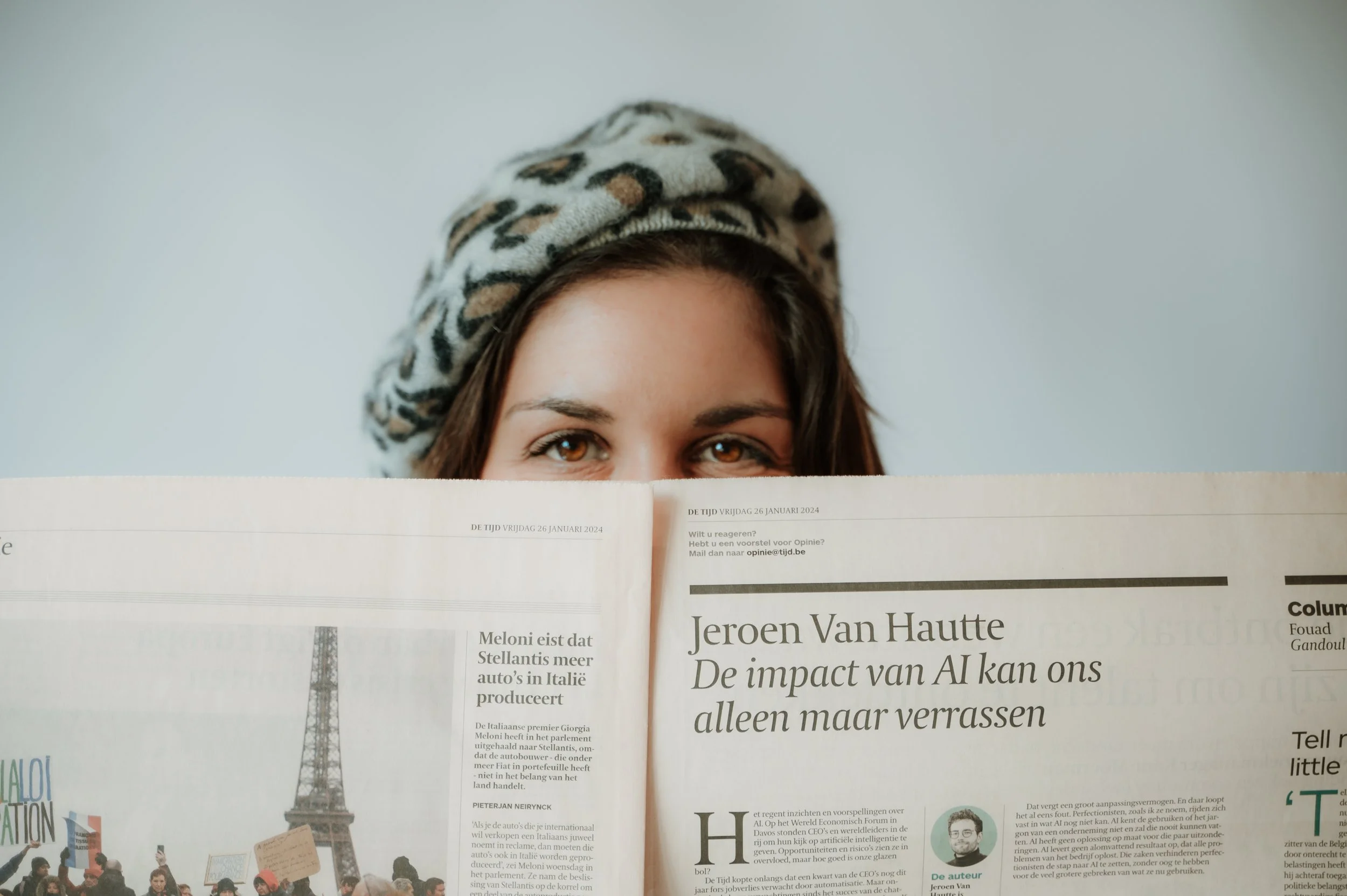 A woman with brown eyes and dark hair partially visible, wearing a leopard print headscarf. She is peeking over a newspaper with a visible Eiffel Tower illustration on the page.
