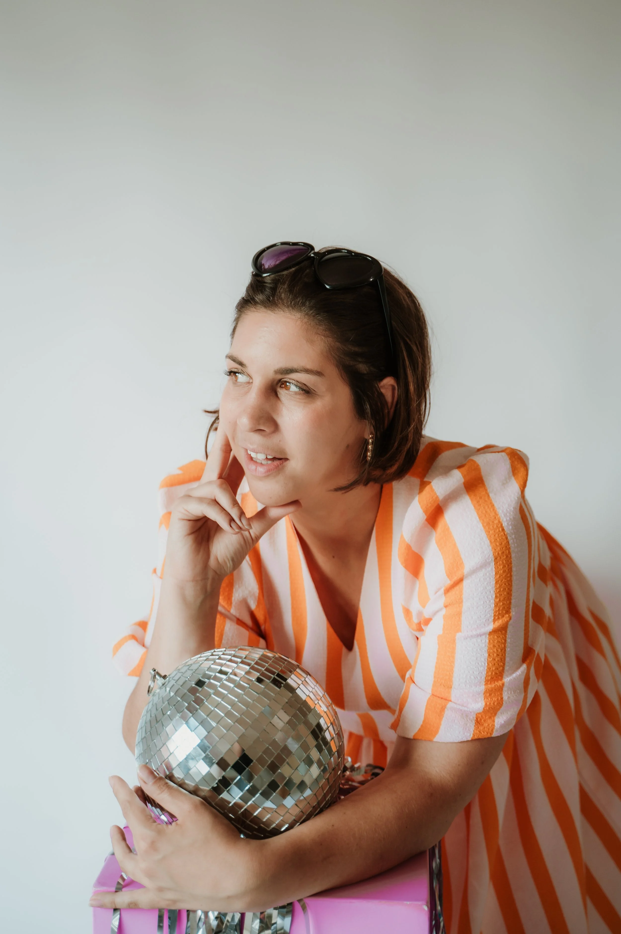 Woman with short brown hair wearing sunglasses on her head, dressed in an orange and white striped shirt, holding a disco ball and a pink gift box.