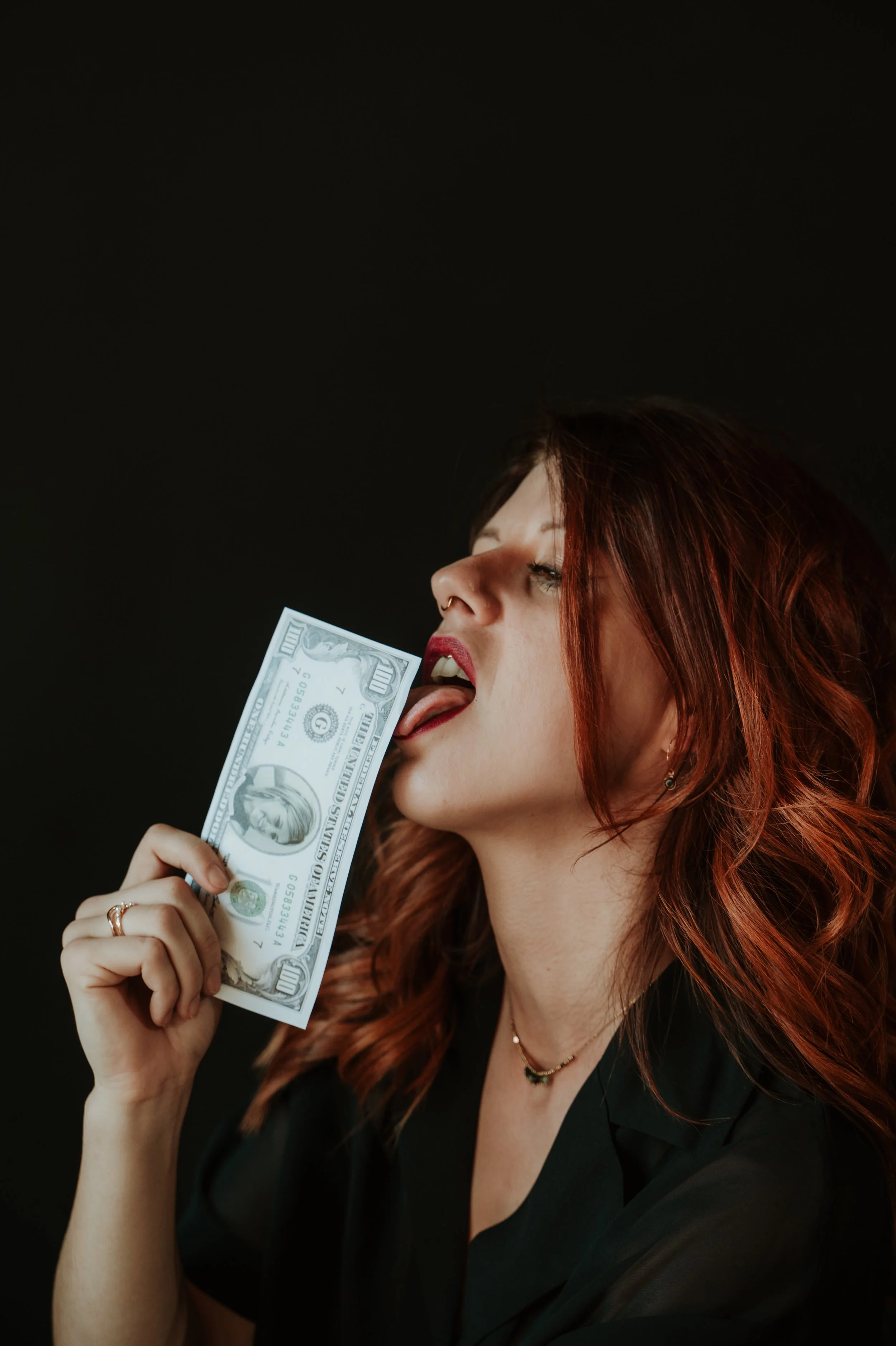 A woman with red hair and makeup licking a hundred-dollar bill against a black background.