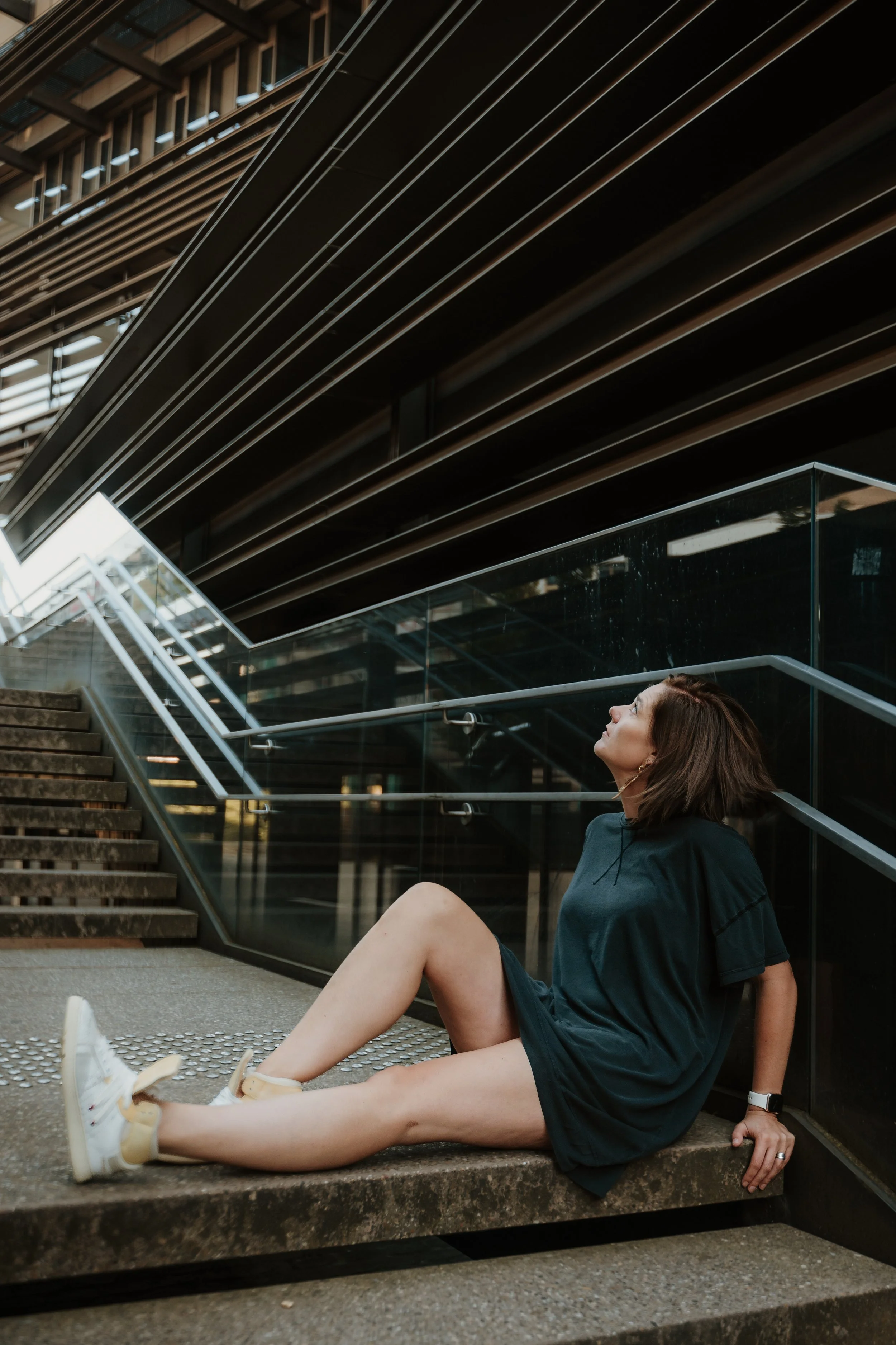 A woman with brown hair sitting on the steps outside a modern building with dark horizontal slats and glass windows. She is wearing a dark t-shirt, shorts, and white sneakers, and is looking upward with a relaxed expression.
