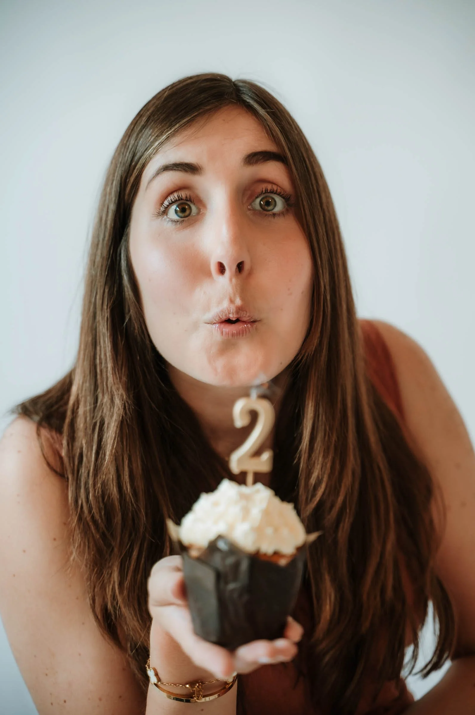 A woman with long brown hair and green eyes holding a chocolate cupcake with white frosting and a '2' candle, wearing a sleeveless top, a bracelet, and a wristwatch, celebrating her second birthday.