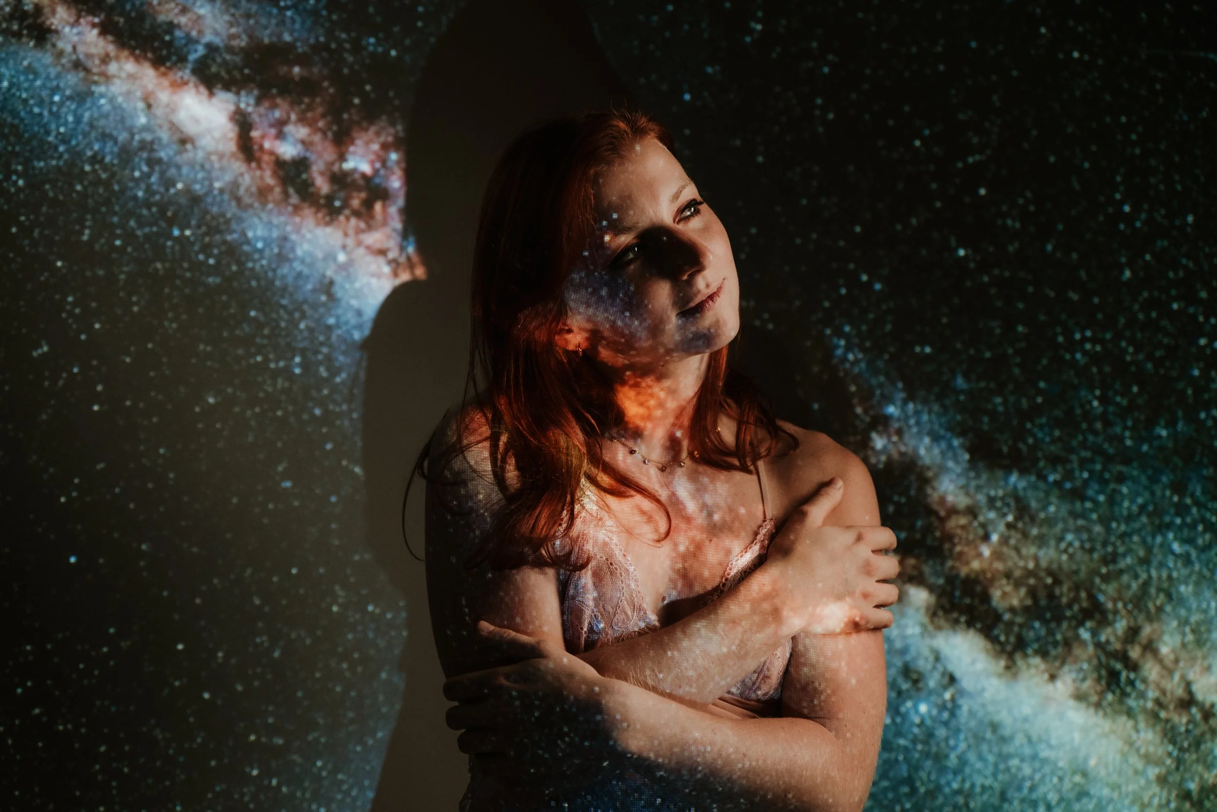 A woman with auburn hair stands with her arms crossed, illuminated by a projection of stars and galaxies on her face and body, creating a cosmic effect.