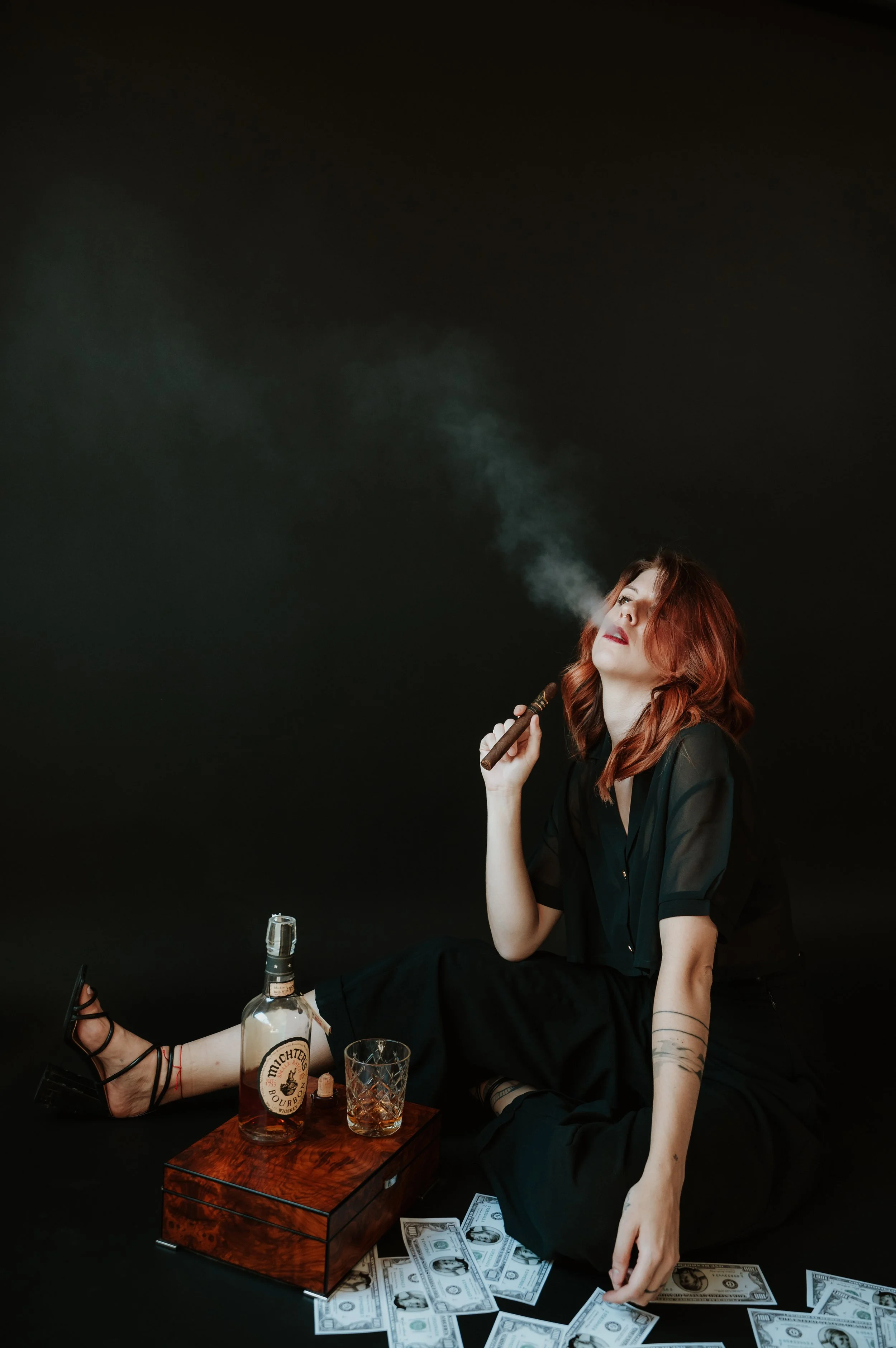 A red-haired woman sitting on the floor wearing black clothes, smoking a cigar, with dollar bills scattered around her and on her lap, and a bottle of liquor on a small wooden table.