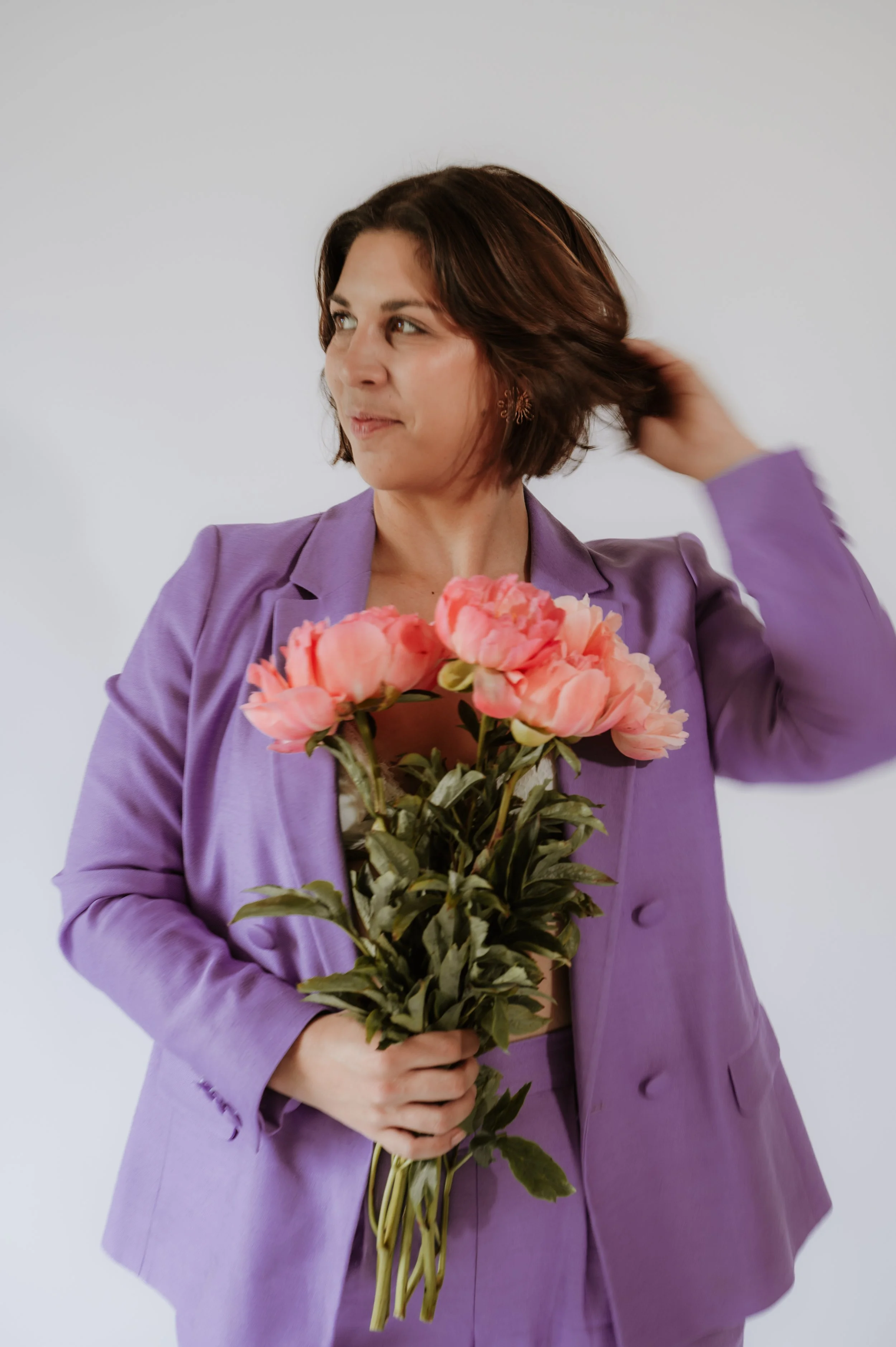 Woman with short brown hair wearing a purple blazer, holding a bouquet of pink flowers, and touching her hair against a plain light gray background.
