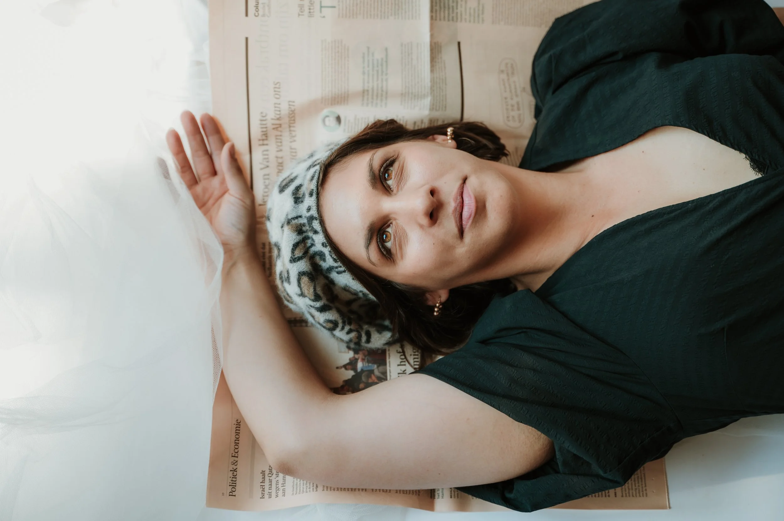 A woman lying on a newspaper, wearing a leopard print headscarf and earrings, looking up with her hand resting on her forehead.