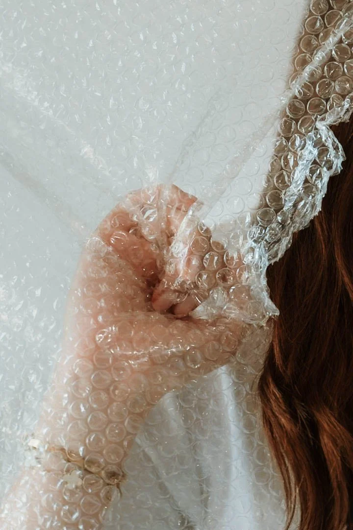 Person's hand holding a sheet of transparent bubble wrap, with part of a white shirt and long brown hair visible.