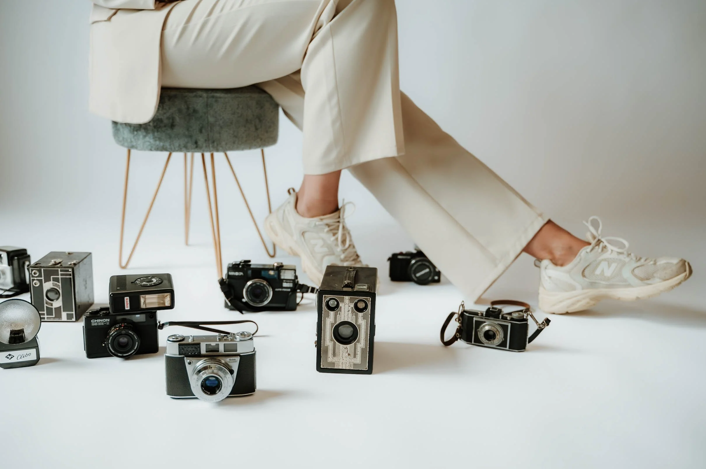 Person sitting on gray velvet chair wearing beige pants and sneakers, surrounded by vintage cameras and camera accessories on white background.