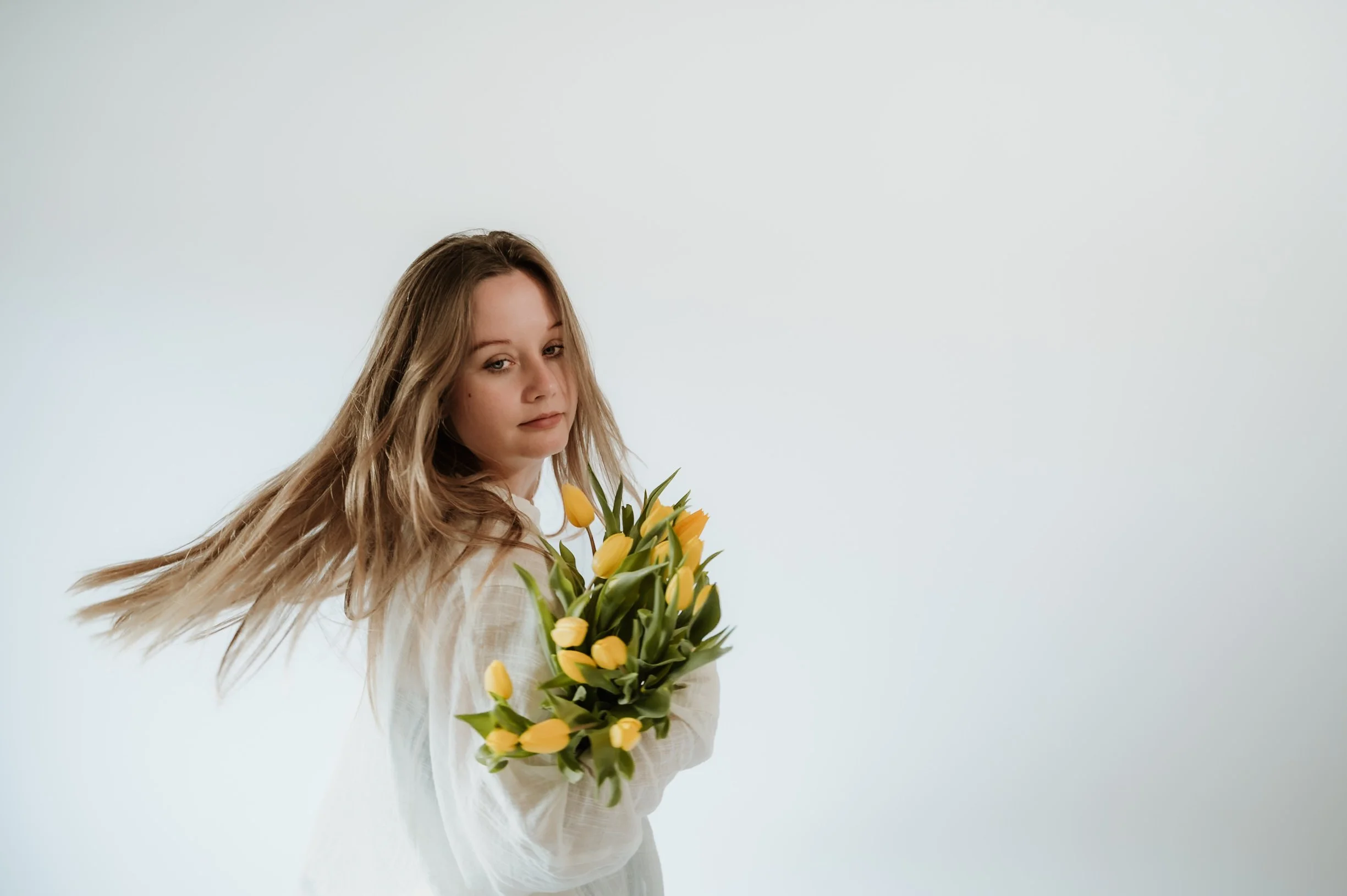 A young woman with long, wavy blonde hair holding a bouquet of yellow tulips, standing against a plain white background.