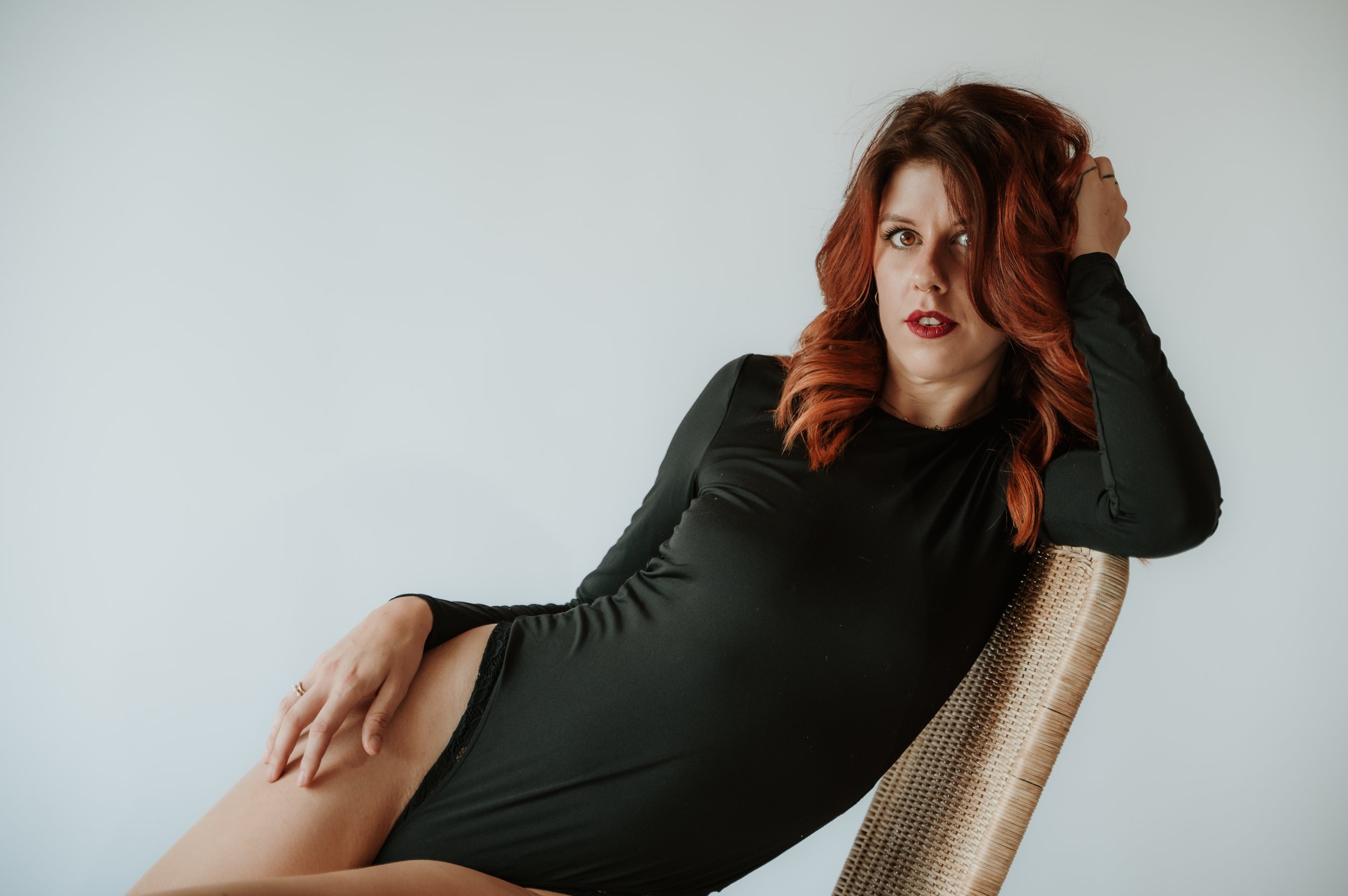 A woman with red hair and light skin, wearing a black long-sleeve dress, sits on a woven rattan chair against a plain light gray background. She is touching her thigh with her left hand and has her right arm raised behind her head, looking at the cam