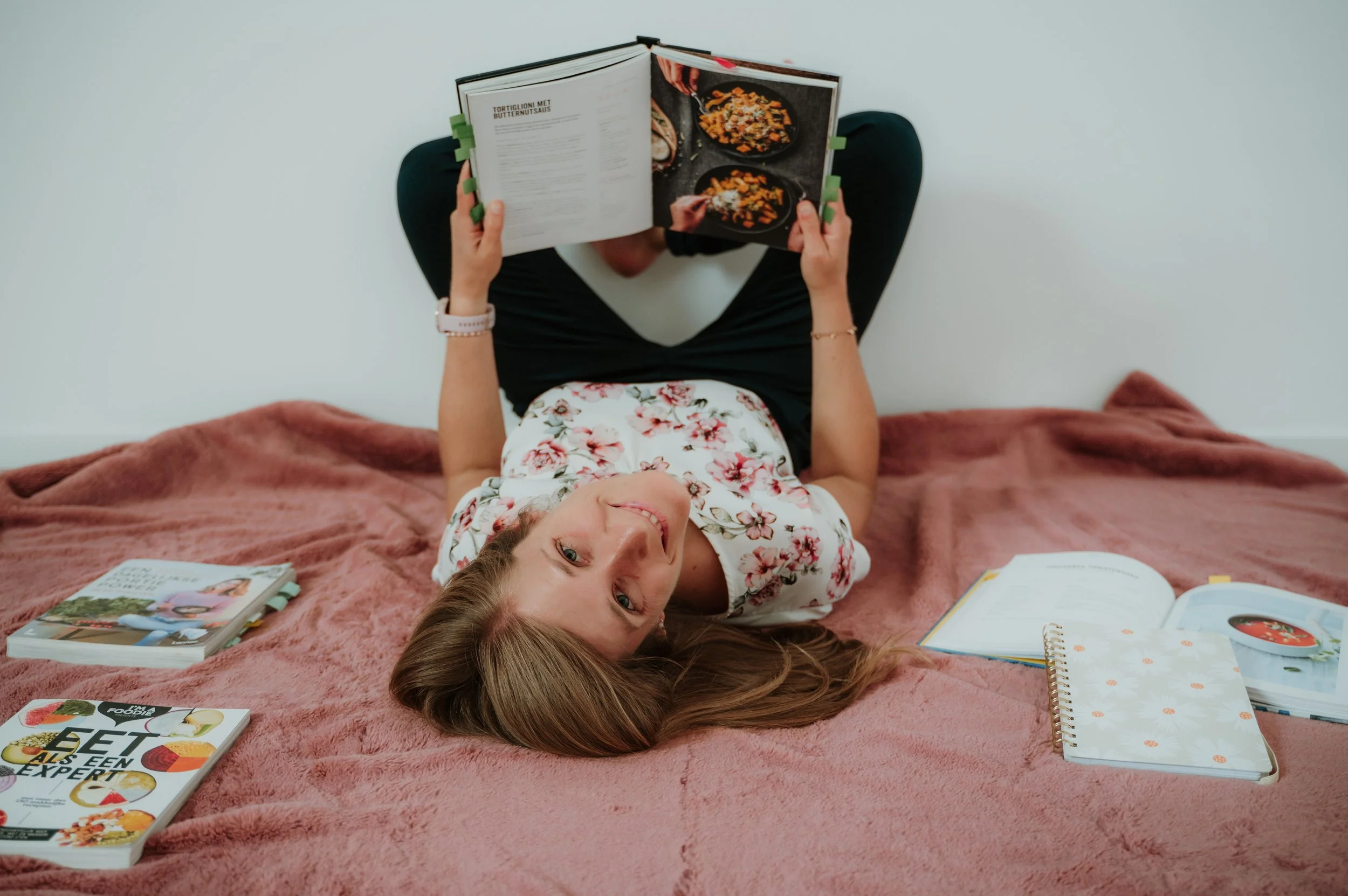 A woman with long brown hair and a floral top is lying on a pink blanket, smiling up at the camera while reading a cookbook. There are magazines and notebooks around her on the blanket.