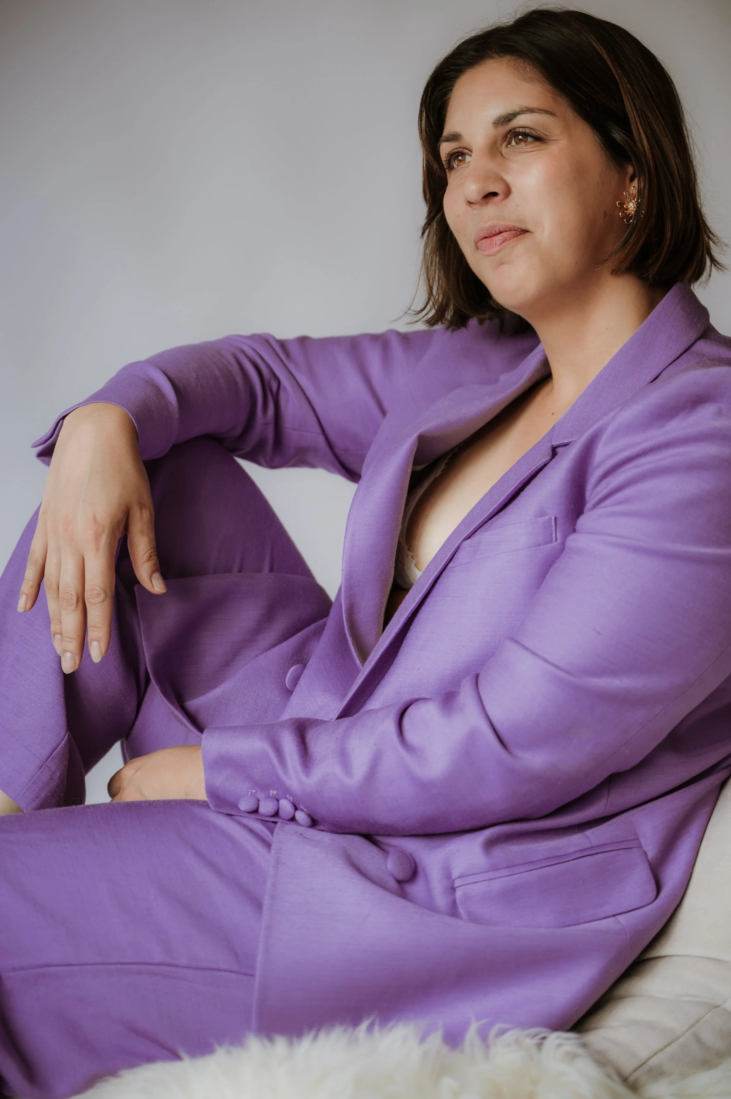 A woman with short brown hair, wearing a purple suit, sitting on a white furry surface against a plain light-colored background, looking thoughtfully into the distance.