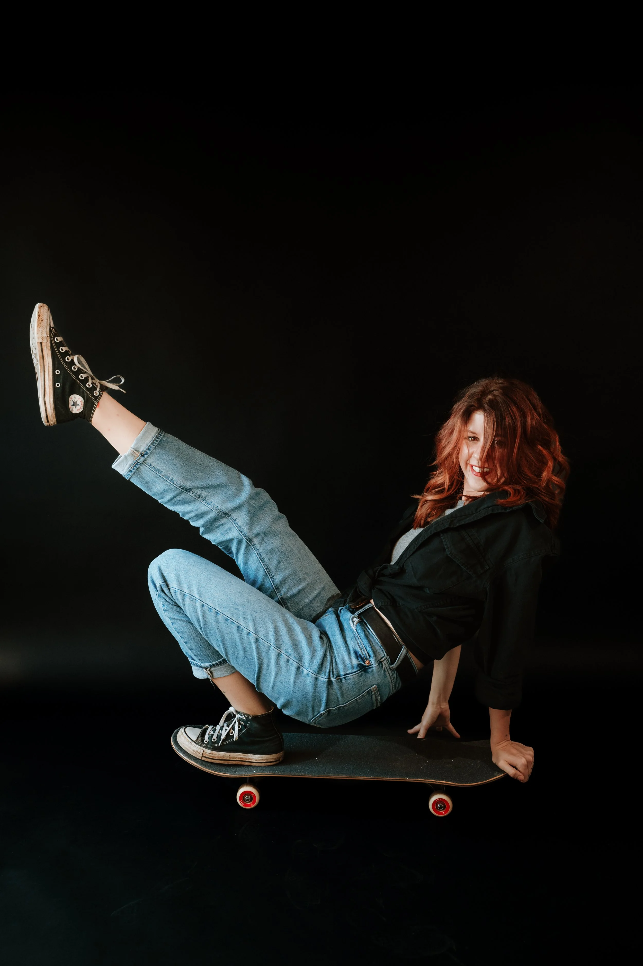 A young woman with curly red hair, wearing a black hoodie and blue jeans, performs a handstand on a skateboard against a black background.
