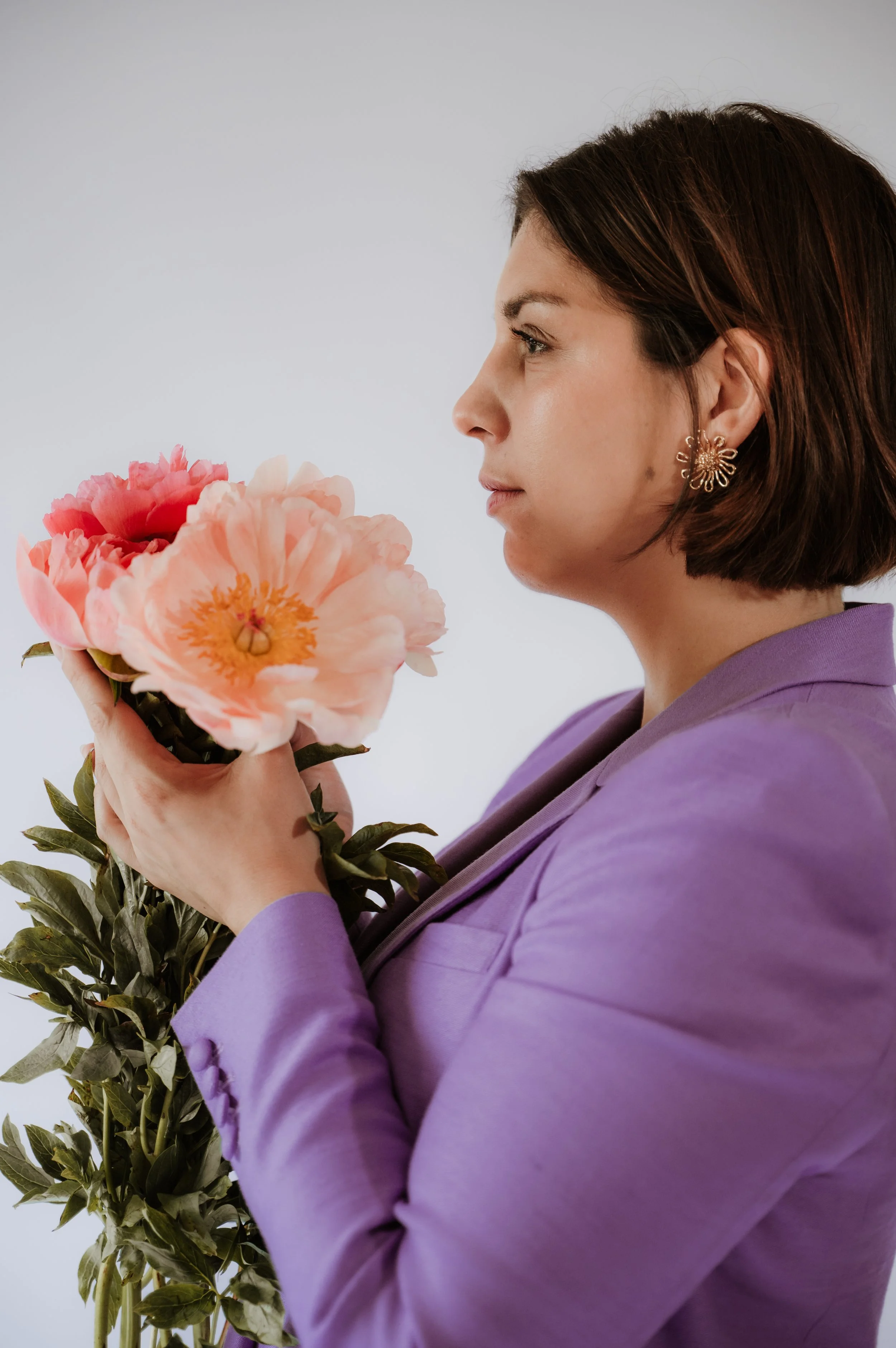 A side profile of a woman with dark brown hair and wearing gold floral earrings, holding a pink peony flower close to her face, against a plain light background, dressed in a lavender blazer.