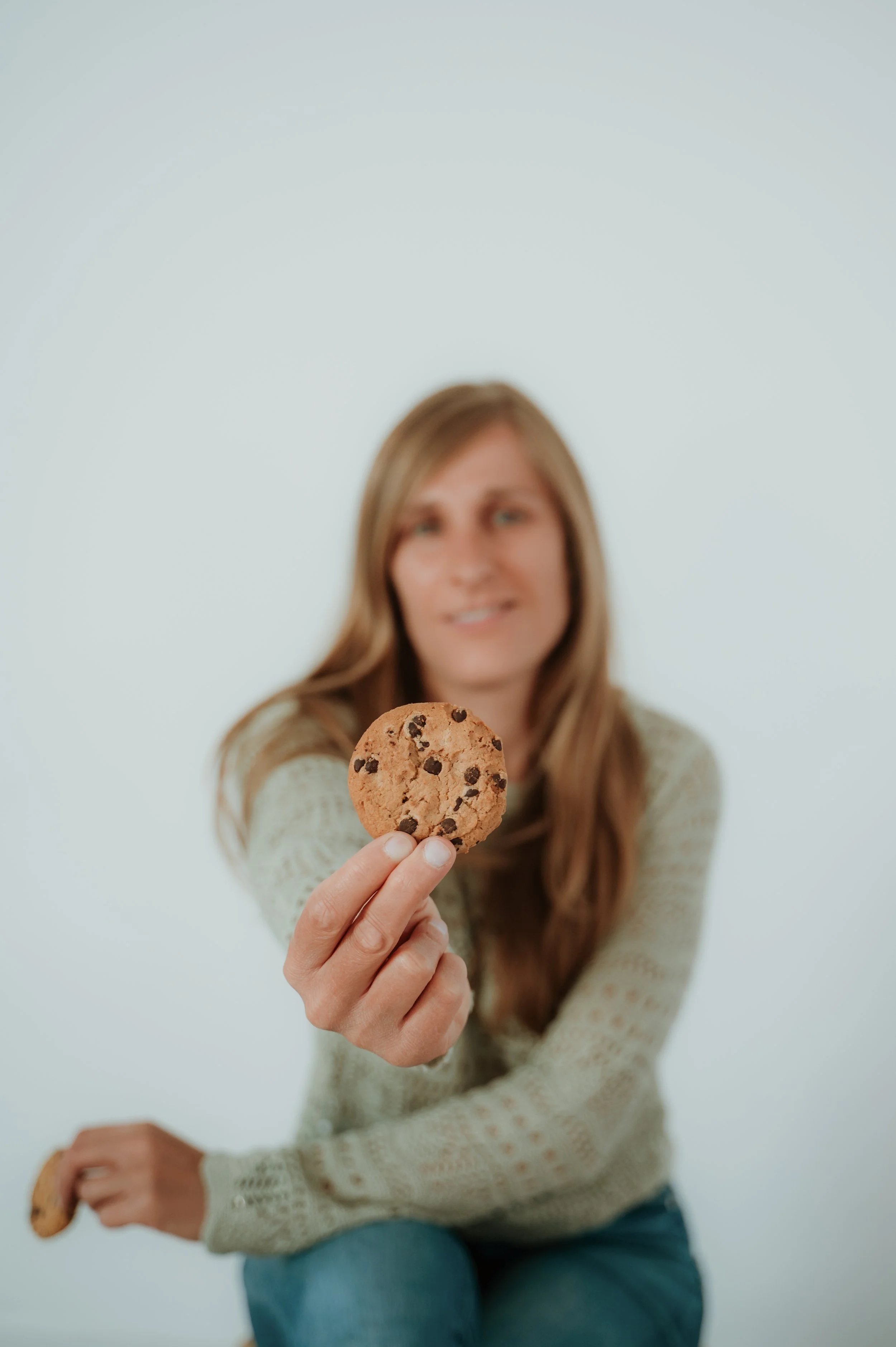 A woman with long brown hair wearing a light-colored sweater, sitting against a plain light background, is holding a chocolate chip cookie close to the camera, with her face slightly out of focus in the background.