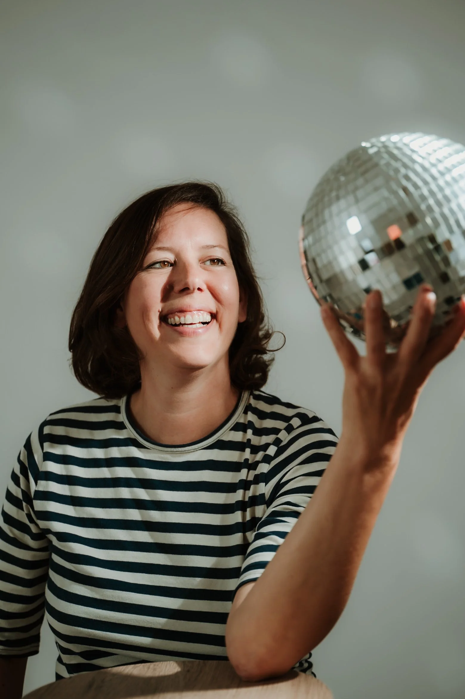 A woman with shoulder-length brown hair wearing a black and white striped shirt, smiling while holding a disco ball in her right hand.