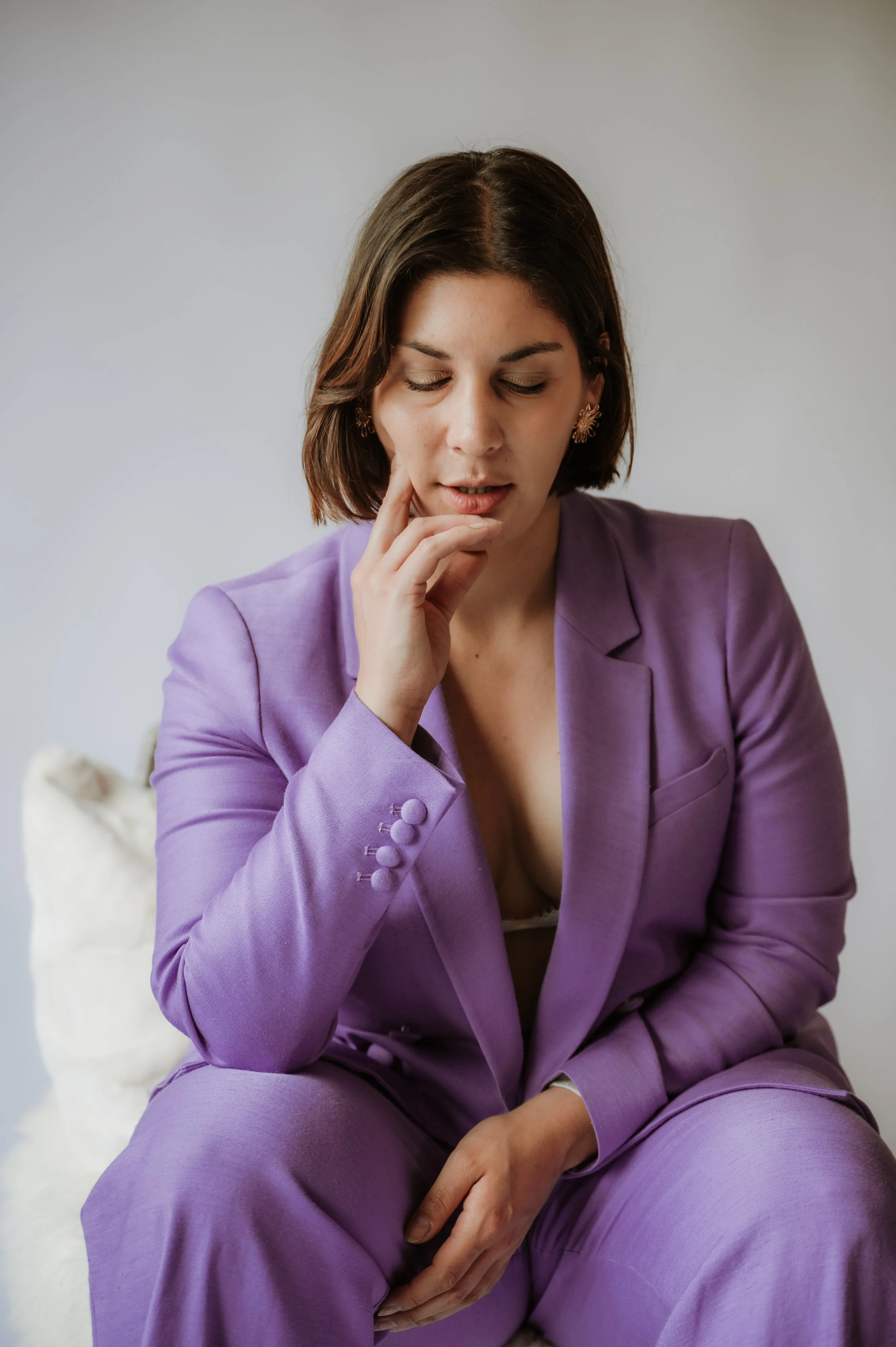 A woman with shoulder-length brown hair, wearing a lavender suit, is sitting with her eyes closed and her hand resting on her chin, appearing thoughtful.