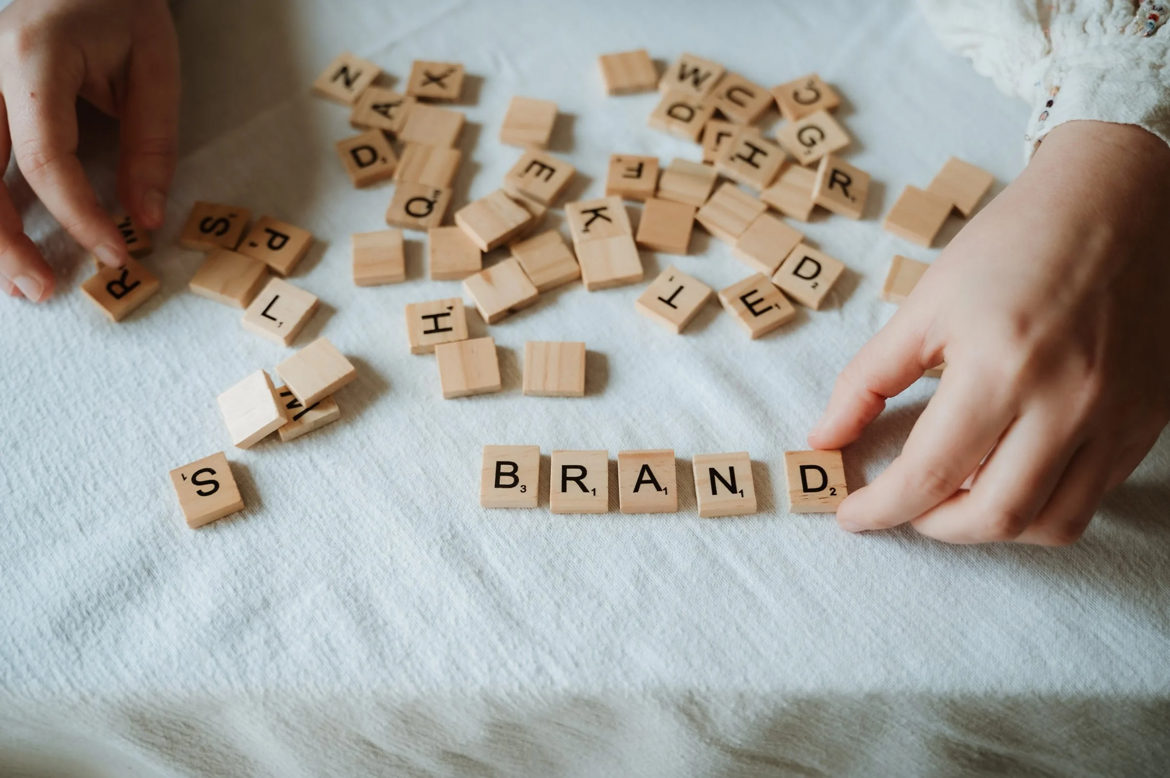 Hands arranging wooden Scrabble tiles spelling 'BRAND' and 'S'.