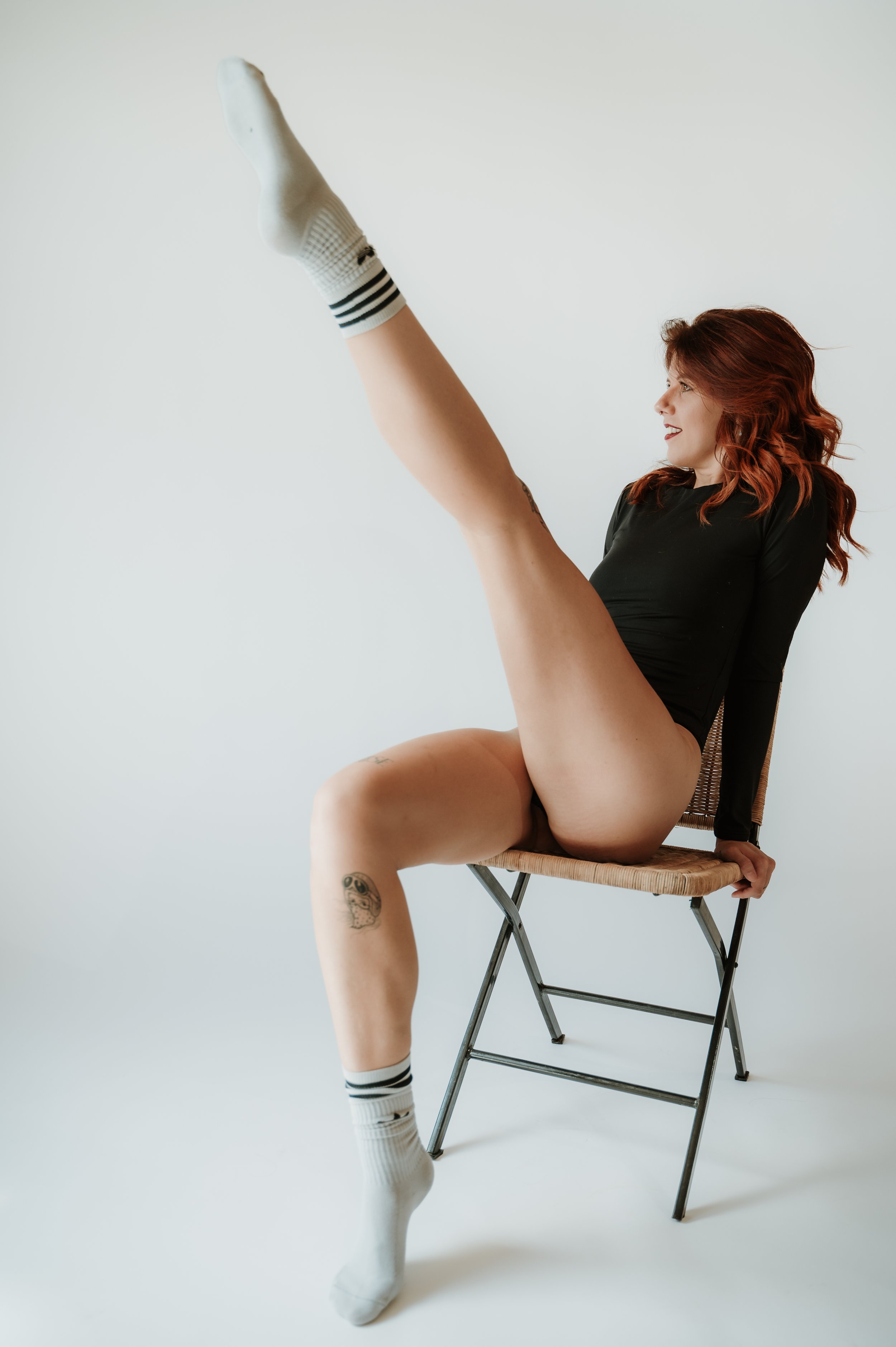A woman with red hair sitting on a chair with her right leg raised high, wearing a black long-sleeve top, white socks with black stripes, and no visible pants, against a plain white background.