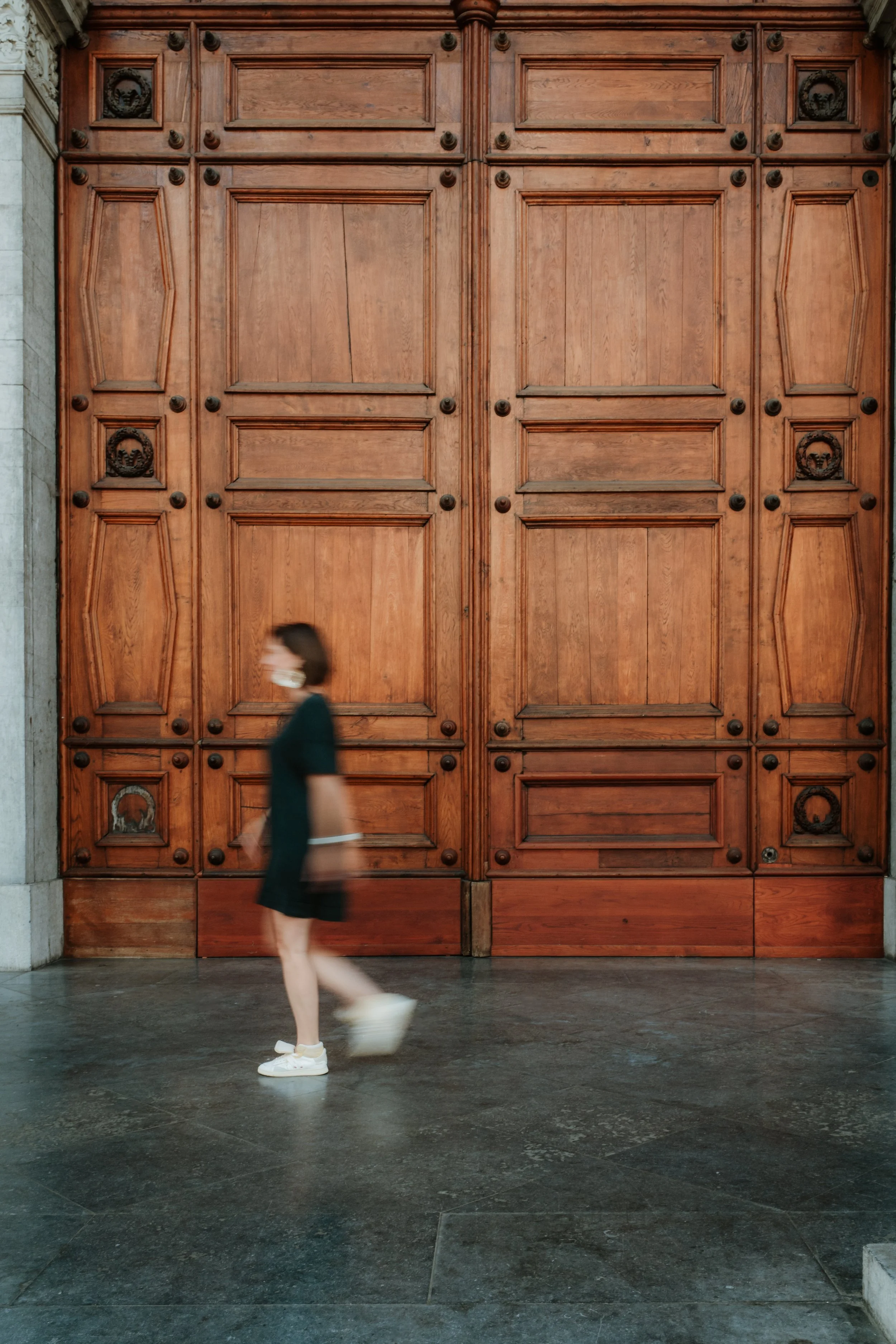 A person walking past a large wooden door with an elaborate design. The person is wearing a dark jacket, shorts, and white sneakers, and is mid-step with motion blur, indicating movement.
