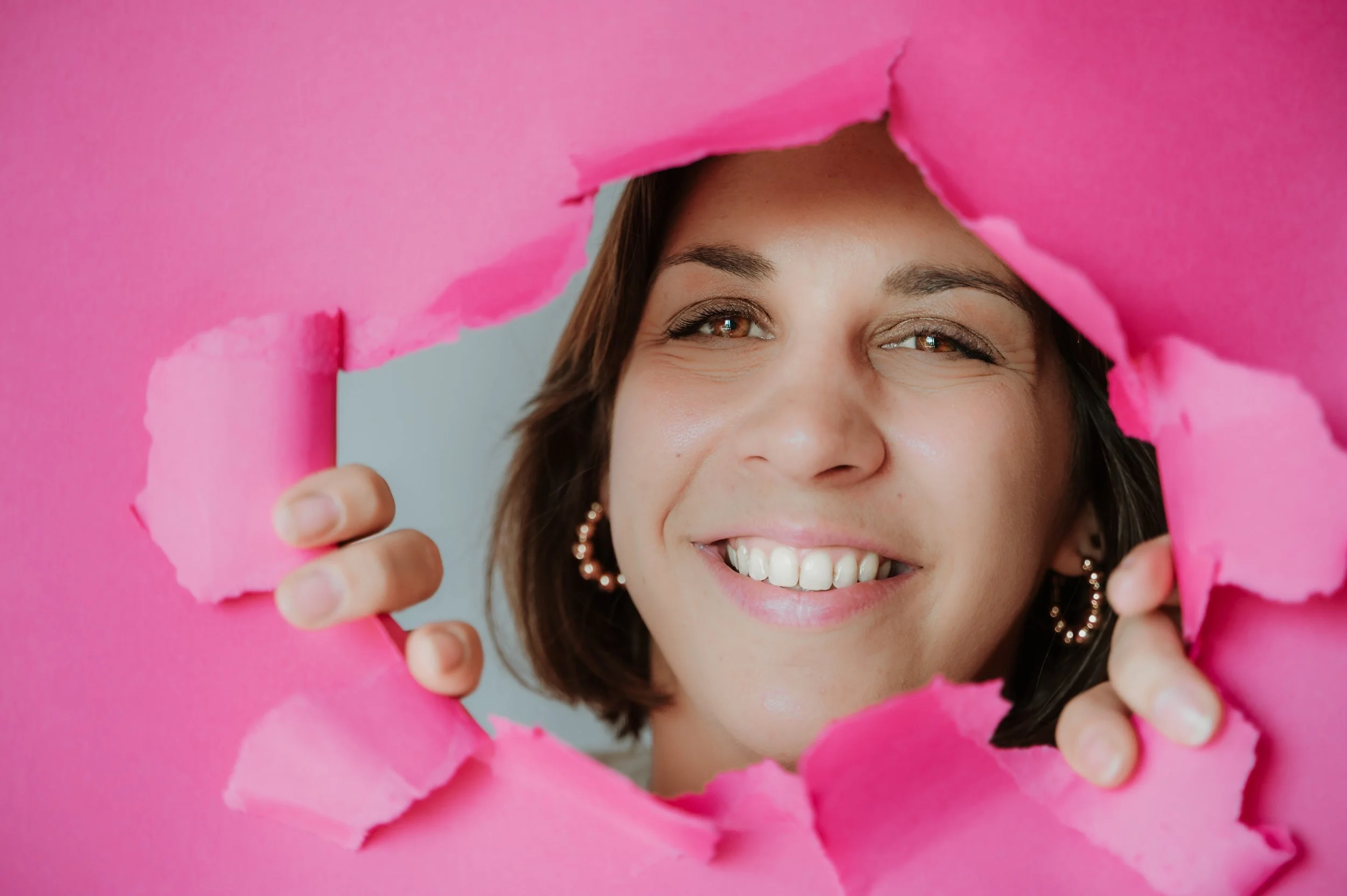 A woman smiling through a torn pink paper background.