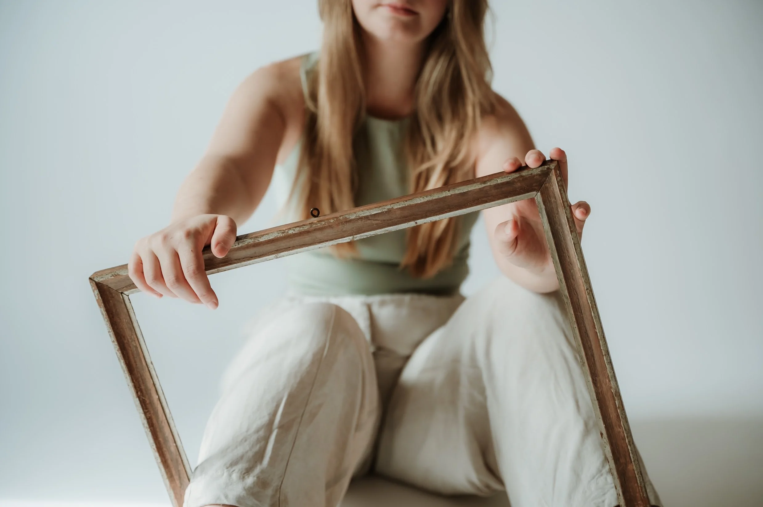 A woman with long, wavy hair holding a weathered wooden picture frame in front of her. She is wearing a light-colored sleeveless top and cream-colored pants, sitting against a plain, light background.