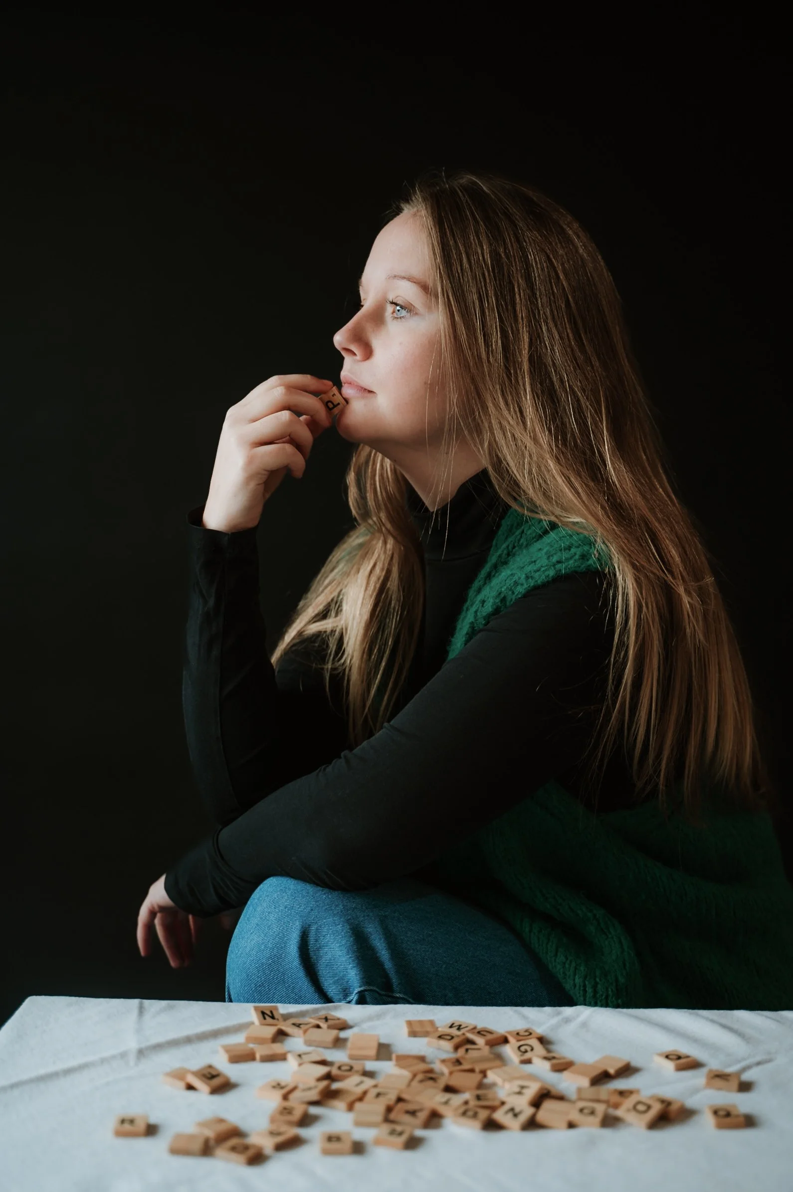 A young woman with long blonde hair sitting against a black background, holding a Scrabble tile near her mouth, with a pile of Scrabble tiles on a white cloth in the foreground.