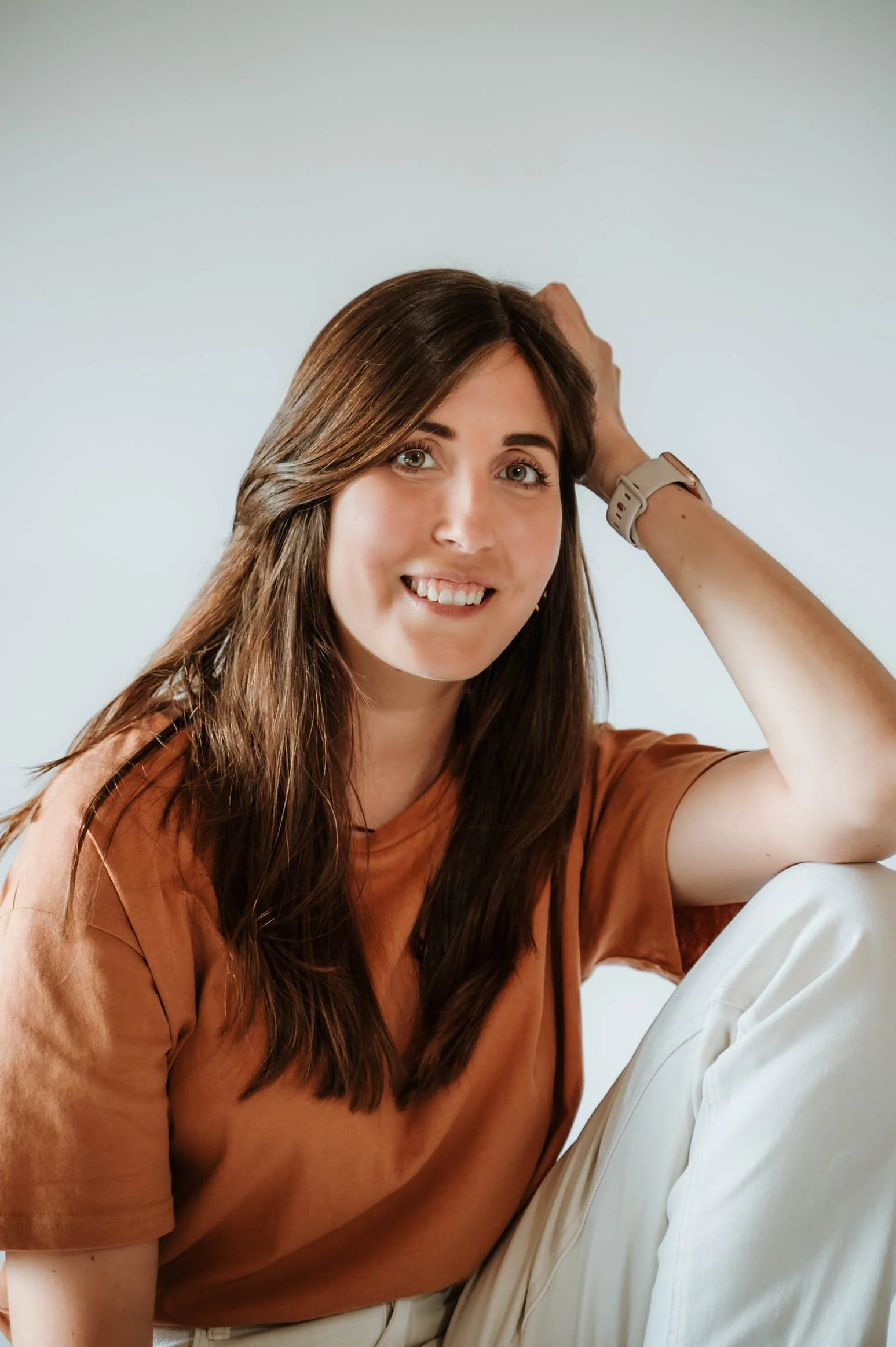 A young woman with long brown hair and green eyes smiling while sitting casually, wearing a tan t-shirt and a smartwatch, against a plain light gray background.