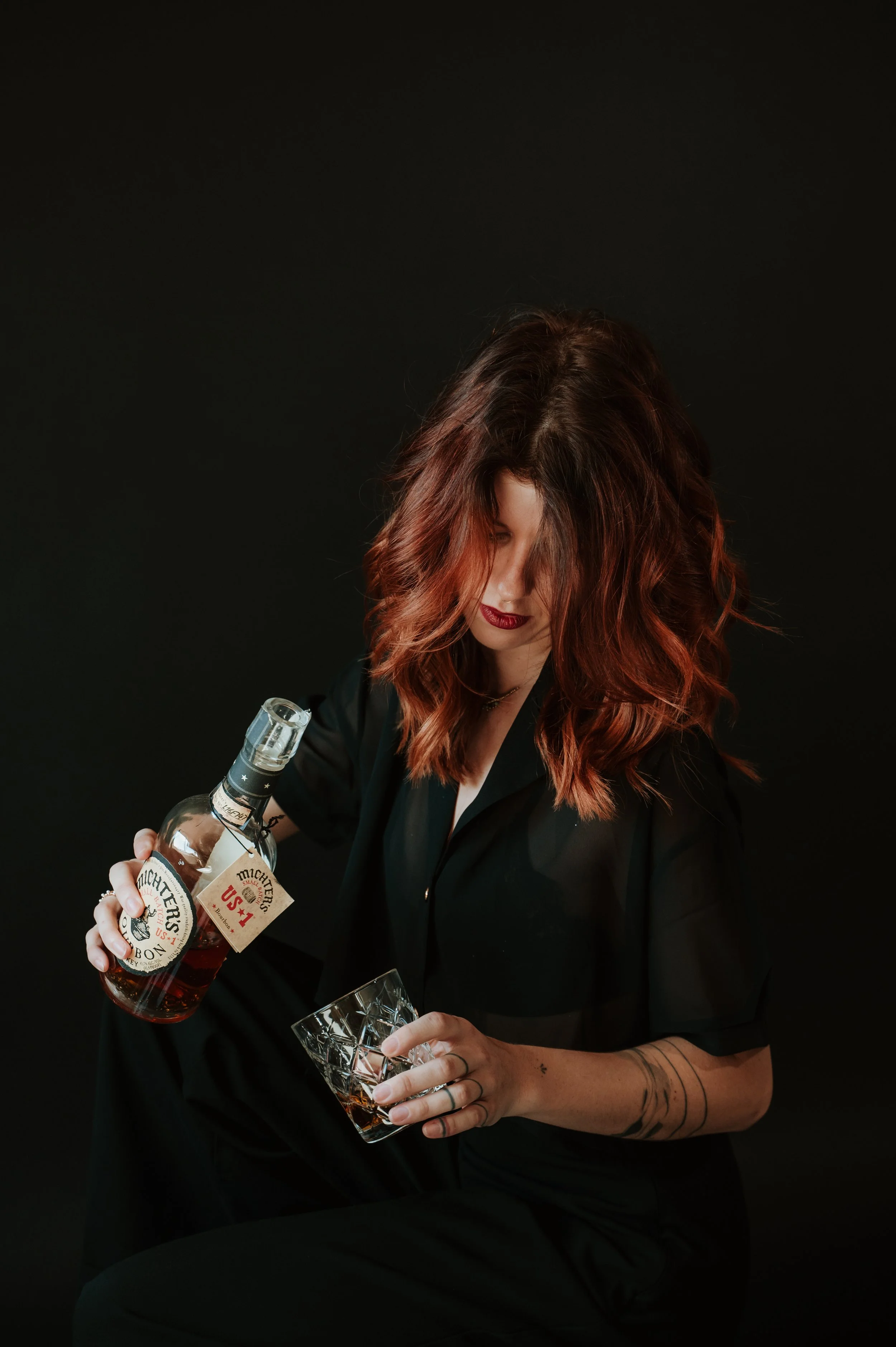 Woman with red hair in black clothing pouring bourbon into a textured glass, against a black background.