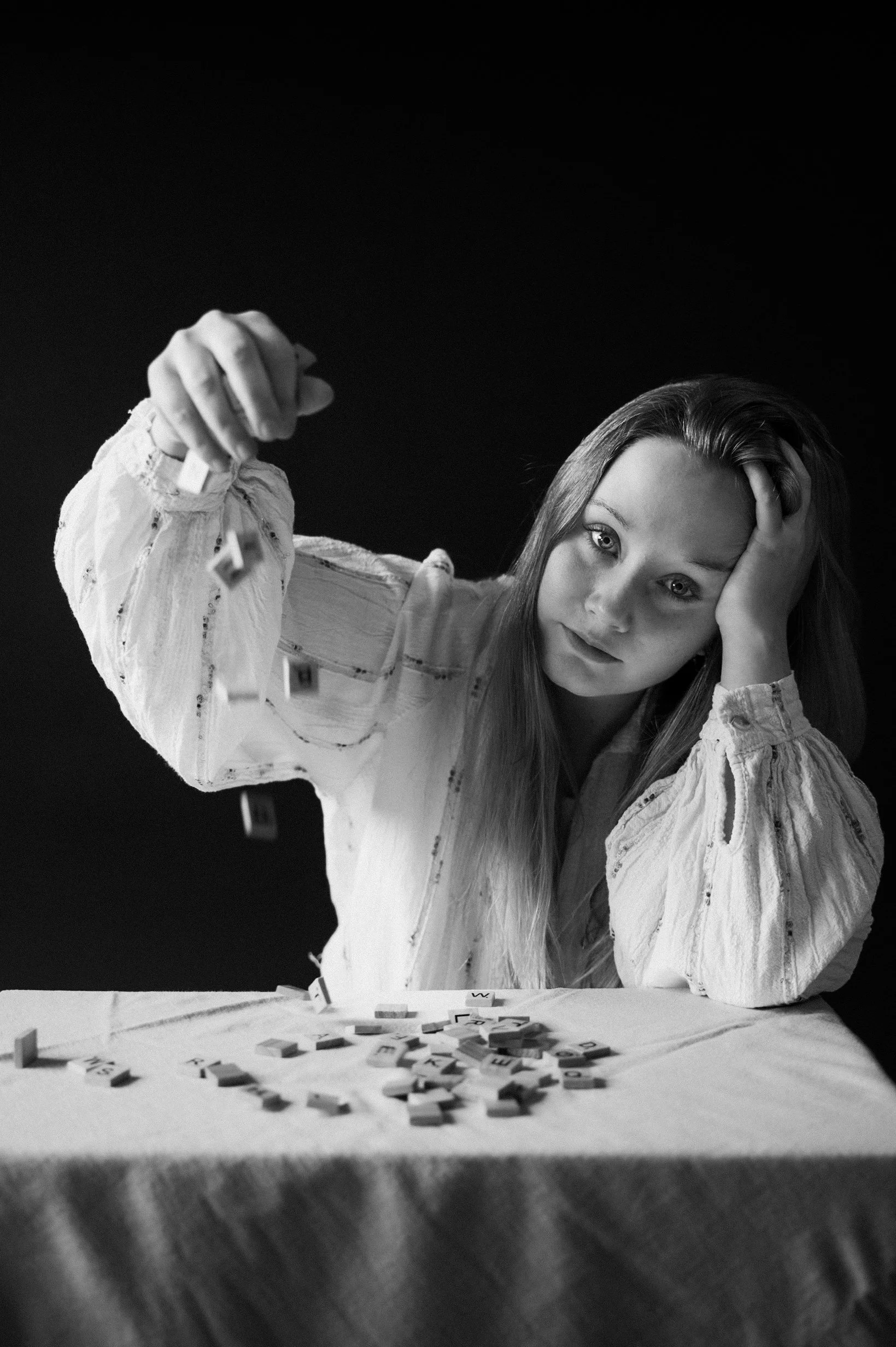 Black and white photo of a young woman with long hair, resting her head on her left hand, looking at the camera, surrounded by scattered letter tiles on a table.