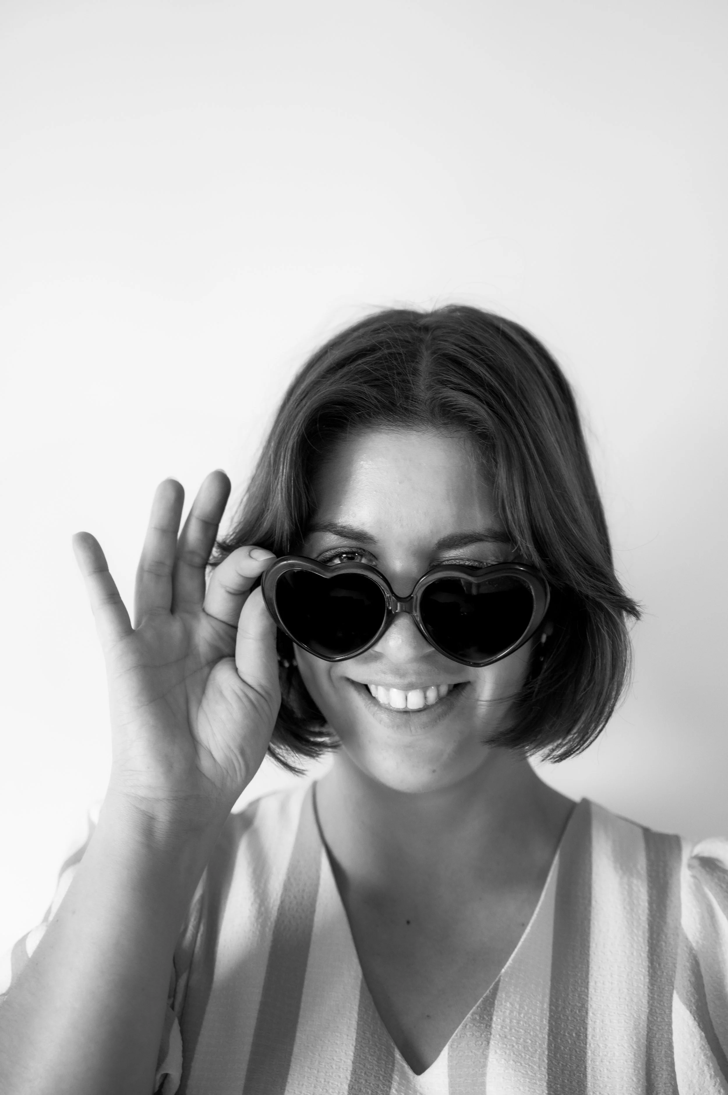 Black and white photo of a young woman with short hair smiling, wearing heart-shaped sunglasses and a sleeveless top with vertical stripes, adjusting her sunglasses with her right hand.