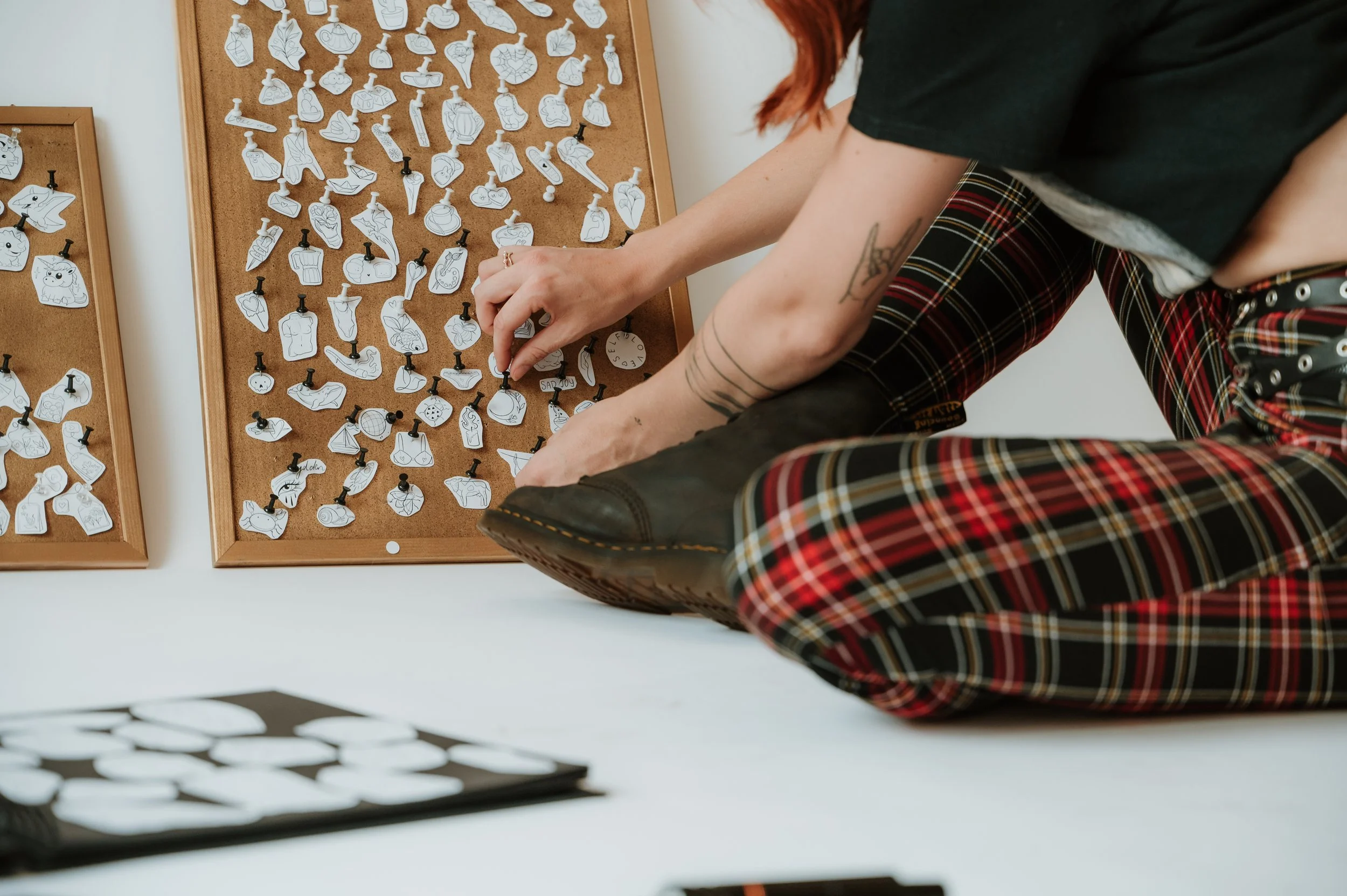 A person with tattoos on their arms is selecting a Halloween-themed pin from a corkboard filled with various pins of different shapes and designs, including pumpkins, ghosts, and other spooky icons.