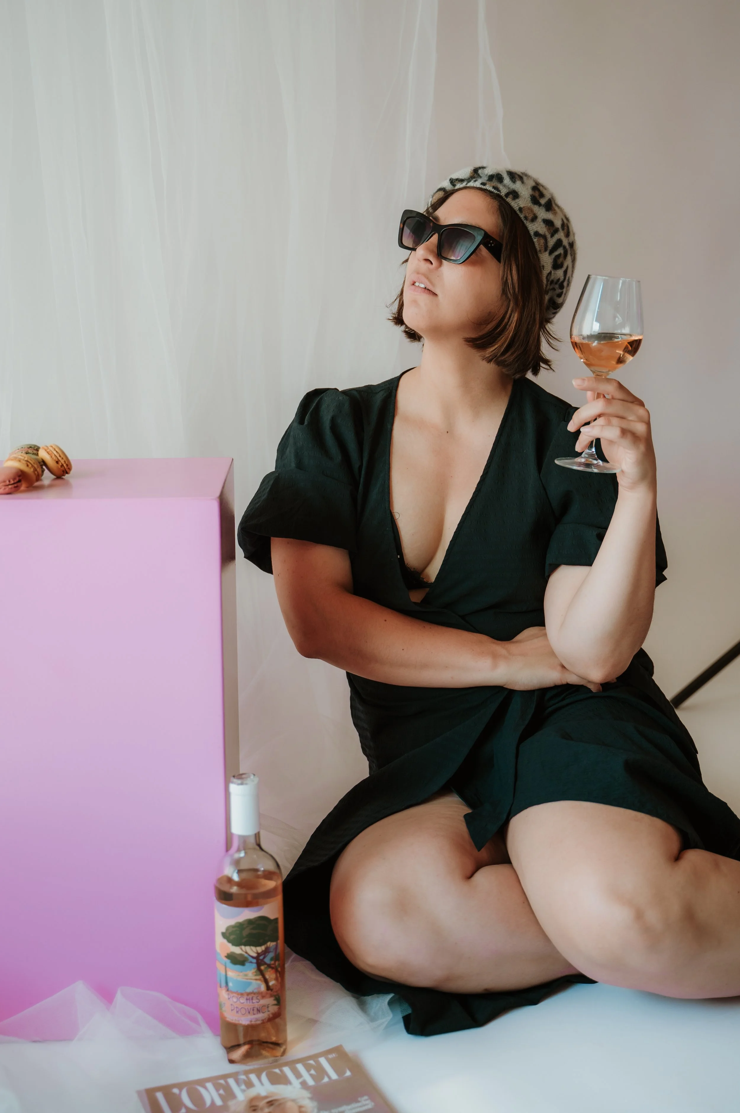 A woman wearing sunglasses, a leopard print beret, and a black dress sitting on the floor, holding a glass of rosé wine. There are macarons on a pink box and a bottle of rose wine nearby. The background is neutral with a sheer fabric draped on the wa