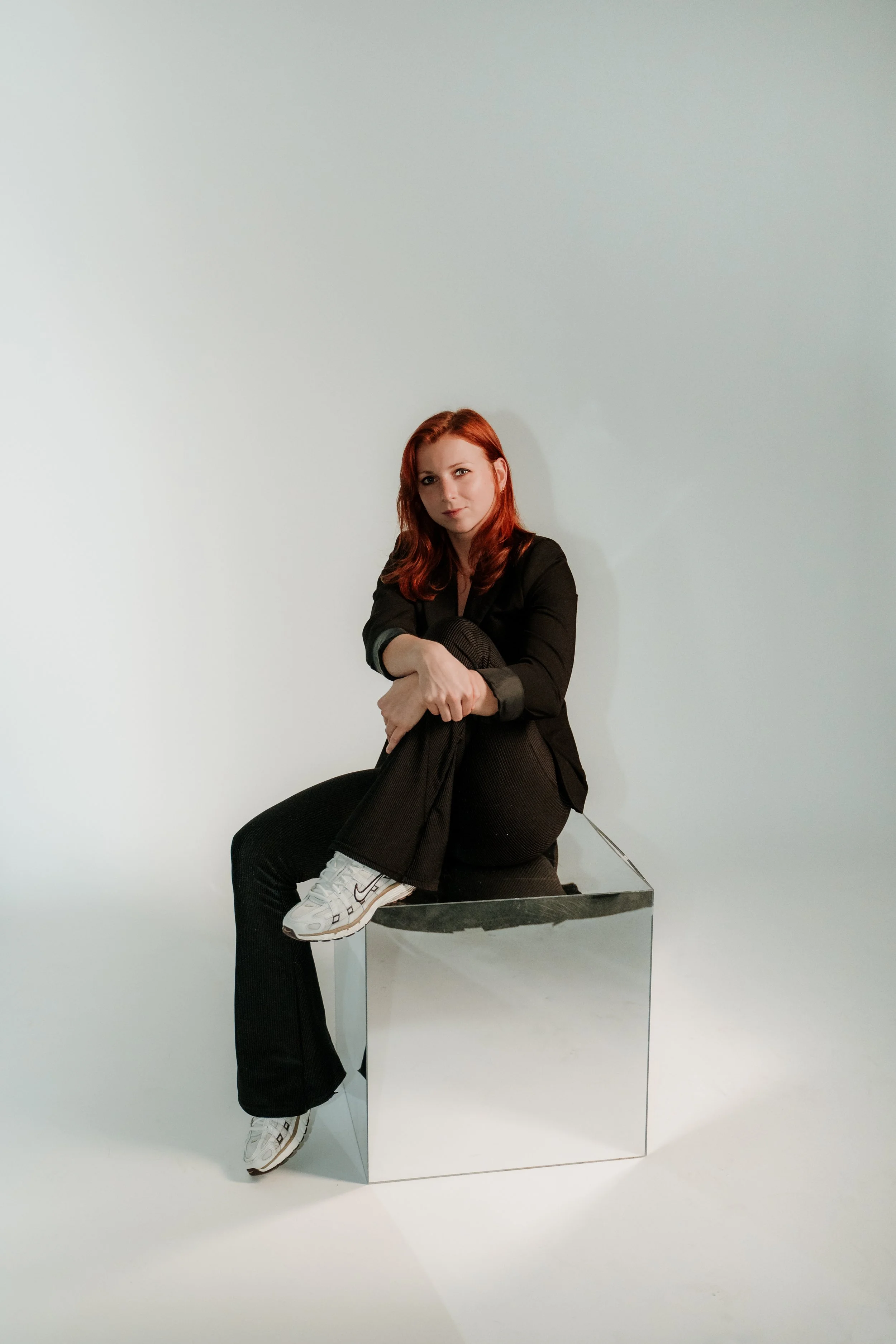 A woman with red hair, wearing a black jacket, seated with her legs crossed on a block in front of a plain light background, looking at the camera.