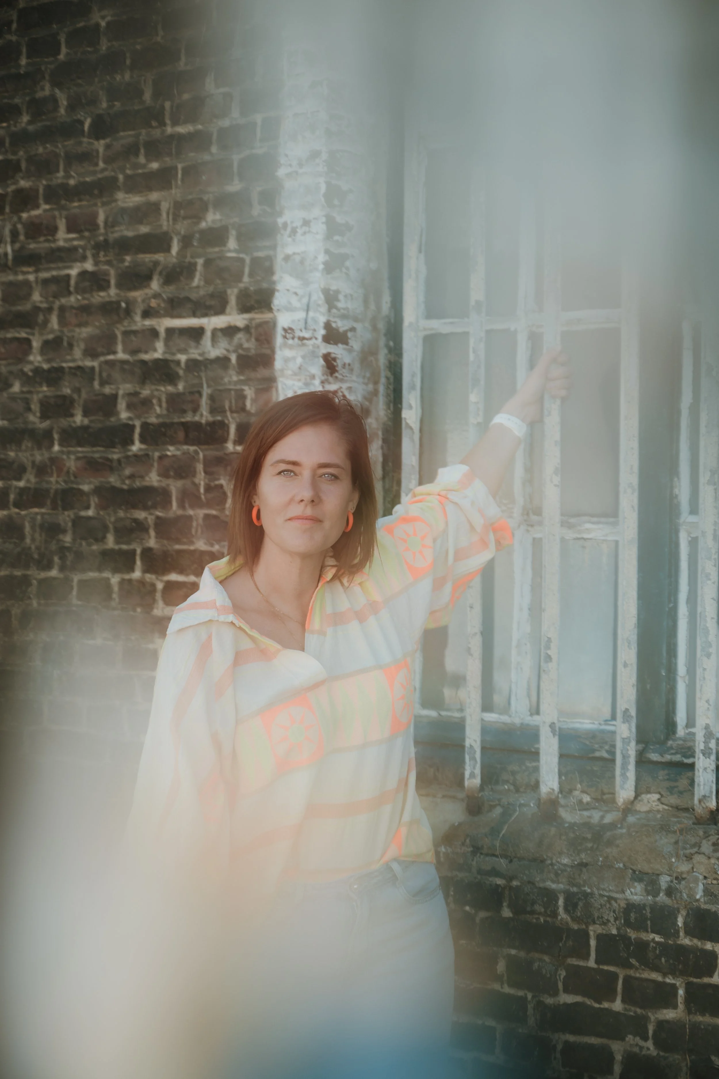 A woman with shoulder-length brown hair, wearing a colorful striped shirt and orange earrings, stands in front of a brick wall and a window frame under construction, pointing at the window frame.