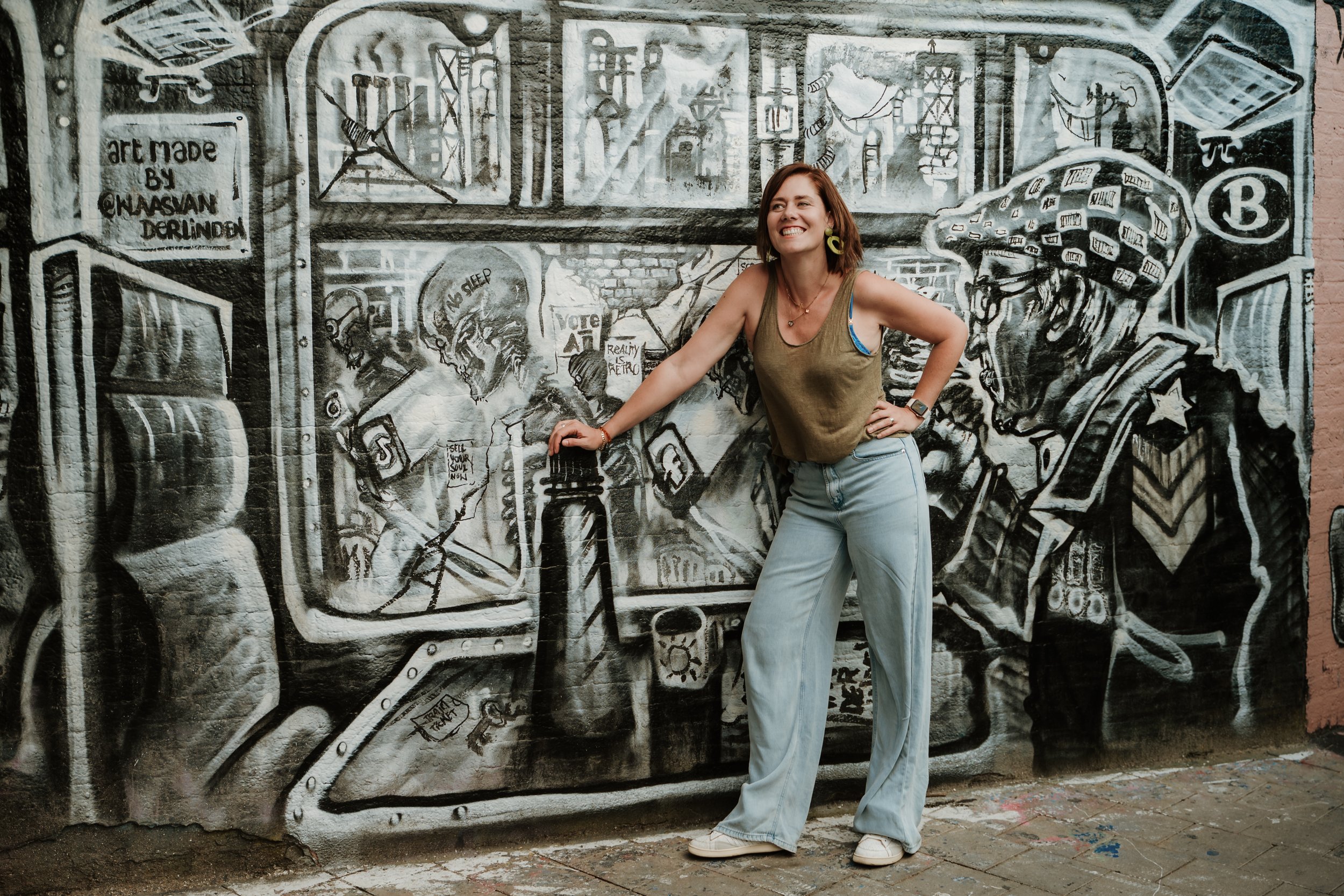 A woman smiling and posing in front of a large black-and-white mural on a brick wall, featuring various images and text such as a skeleton, a vending machine, and artistic sketches.