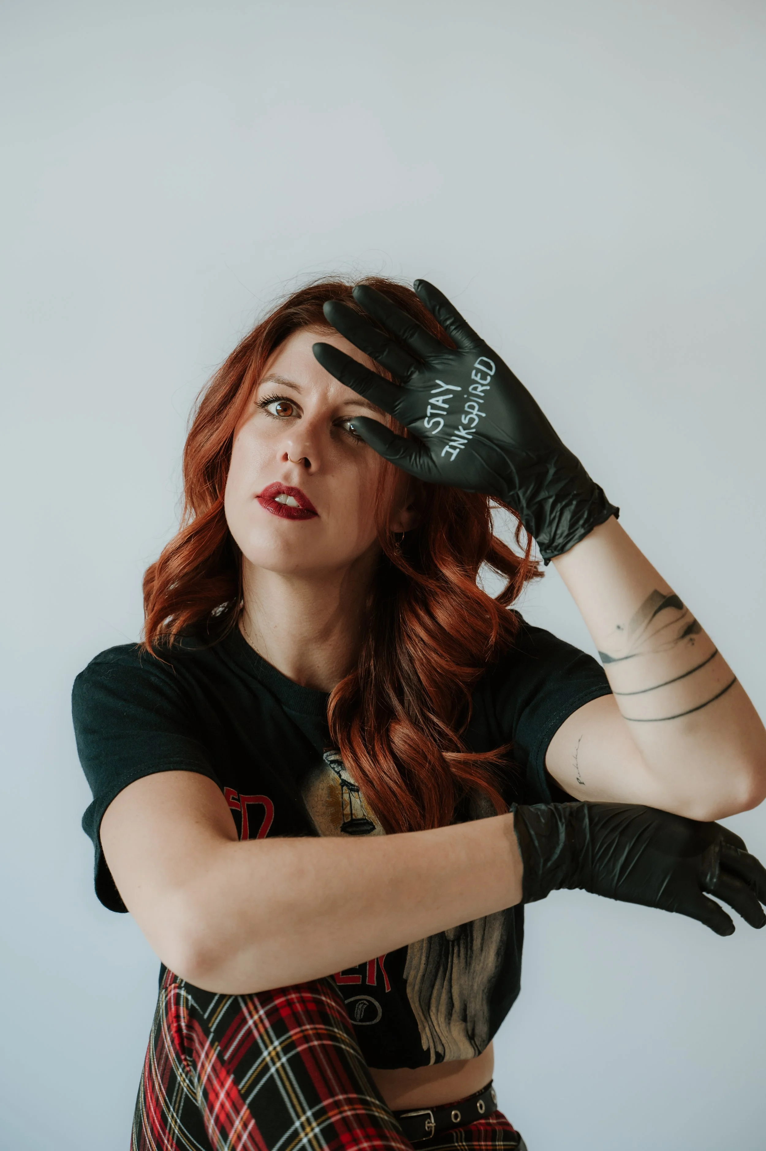 A woman with red hair and tattoos is posing against a plain white background. She wears black gloves with the words "STAY INKSPIRIED" written on one and has a confident expression.