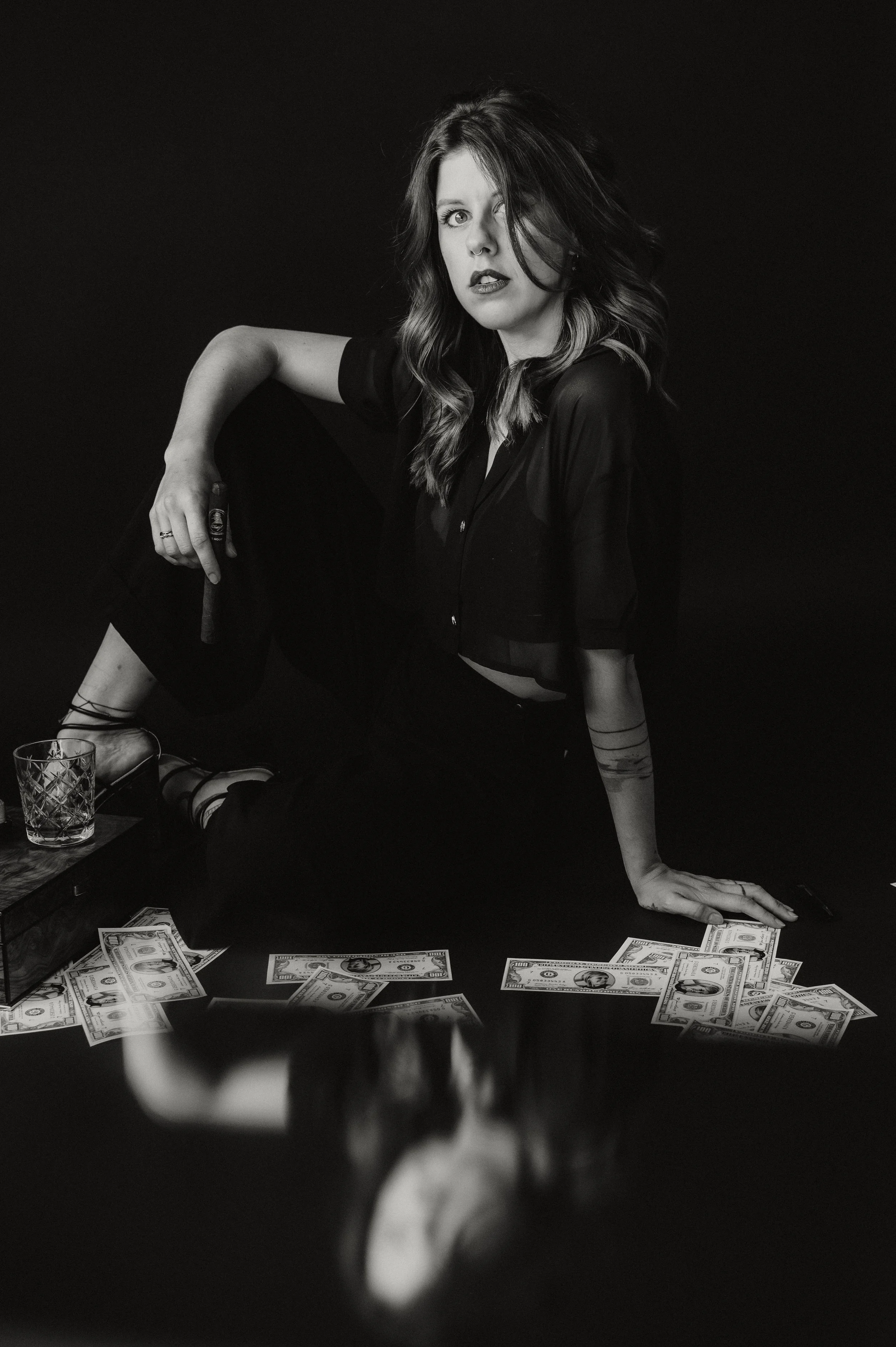 A woman with wavy hair and dark lipstick is sitting on a table surrounded by scattered US dollar bills. She is wearing a dark outfit and has one hand on the table and the other resting on her knee. Various objects, including a glass and money, are in