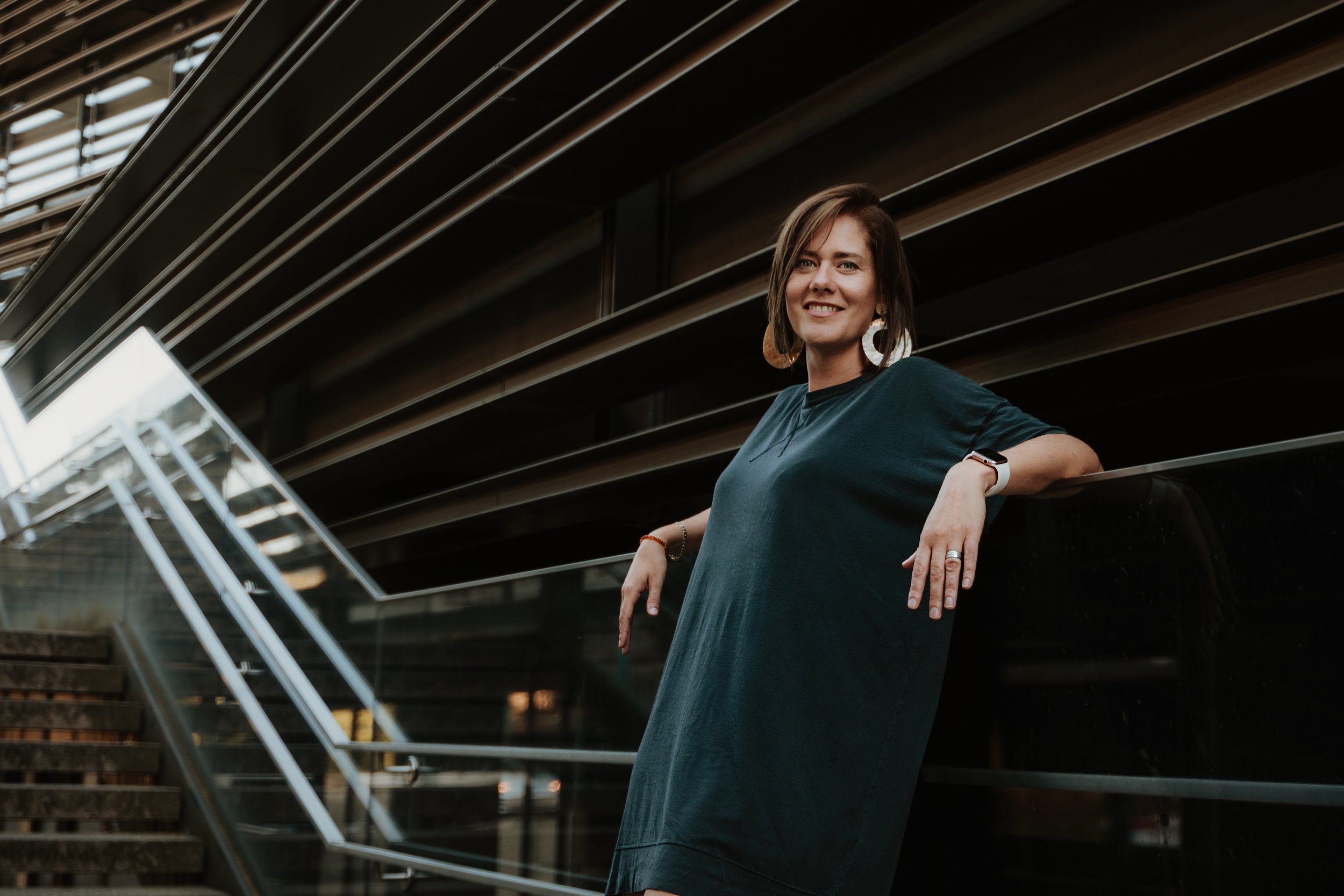 A woman with short brown hair smiling, wearing large earrings and a dark green dress, leaning on a railing in a modern building with horizontal architectural lines.