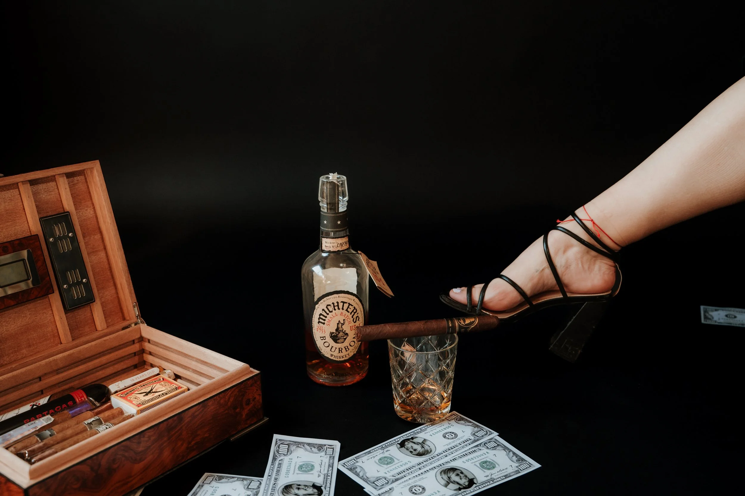 A wooden cigar box, several hundred dollar bills, a bottle of Maker's Mark bourbon whiskey, a glass with a patterned surface, and a woman's leg wearing black high heel shoes and red and black ankle straps against a black background.
