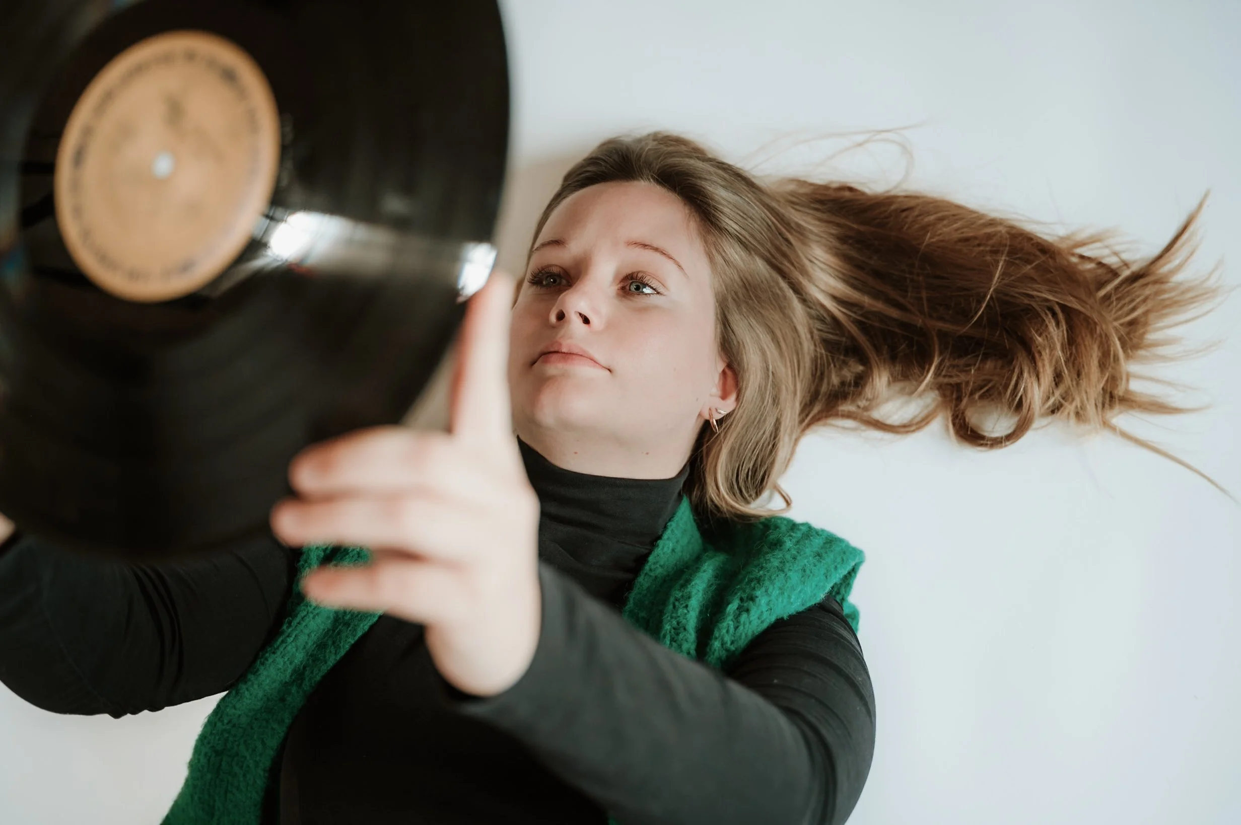 Young woman with long blonde hair taking a selfie with a smartphone, wearing a black shirt and green sweater.