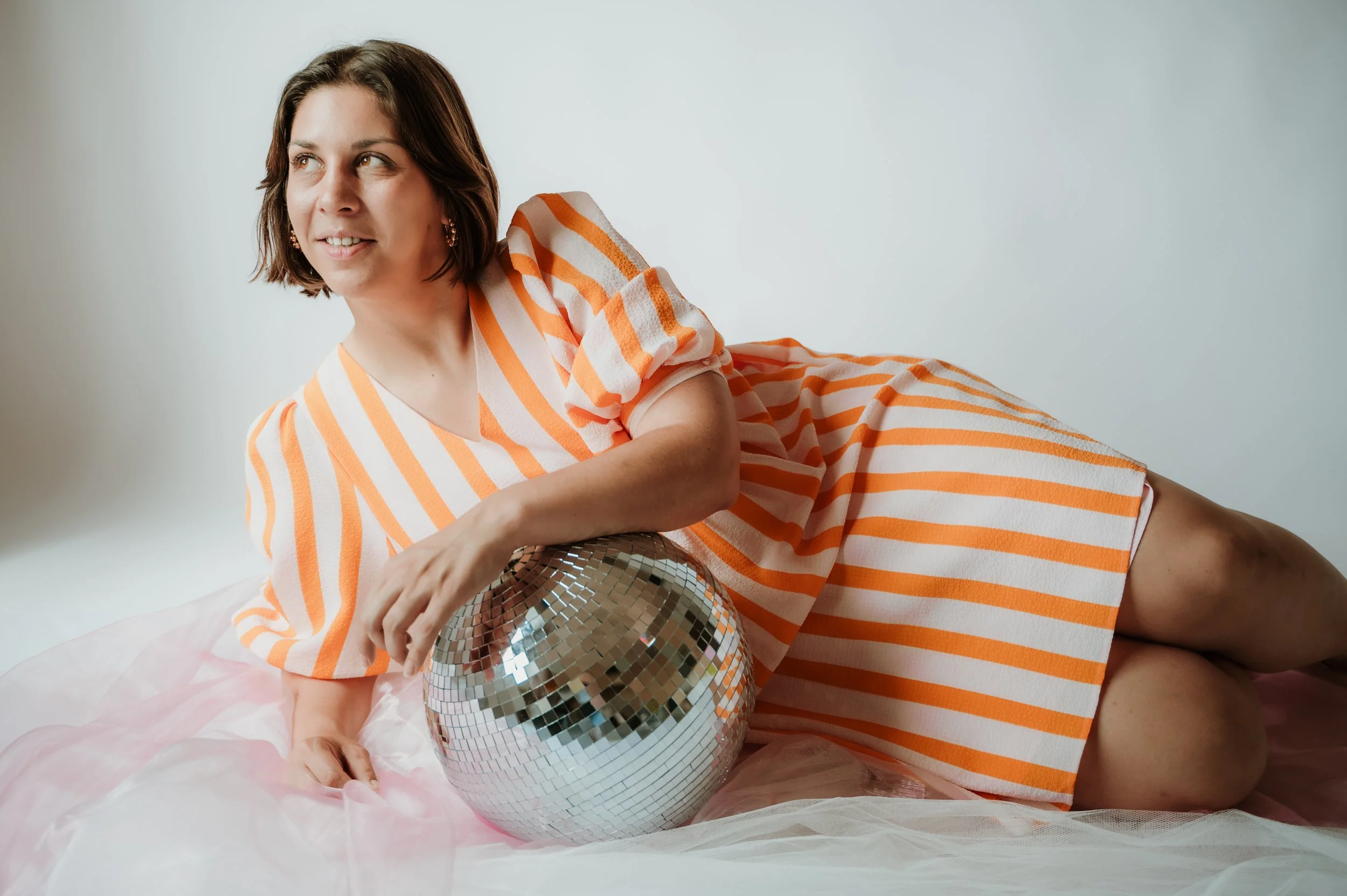 Woman with shoulder-length brown hair wearing an orange and white striped dress, leaning on a disco ball placed on pink tulle fabric, against a plain light background.